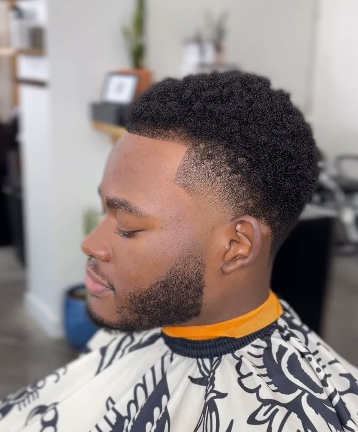 Profile view of a man with a short curly fade and trimmed beard, seated in a barber chair wearing a patterned cape.