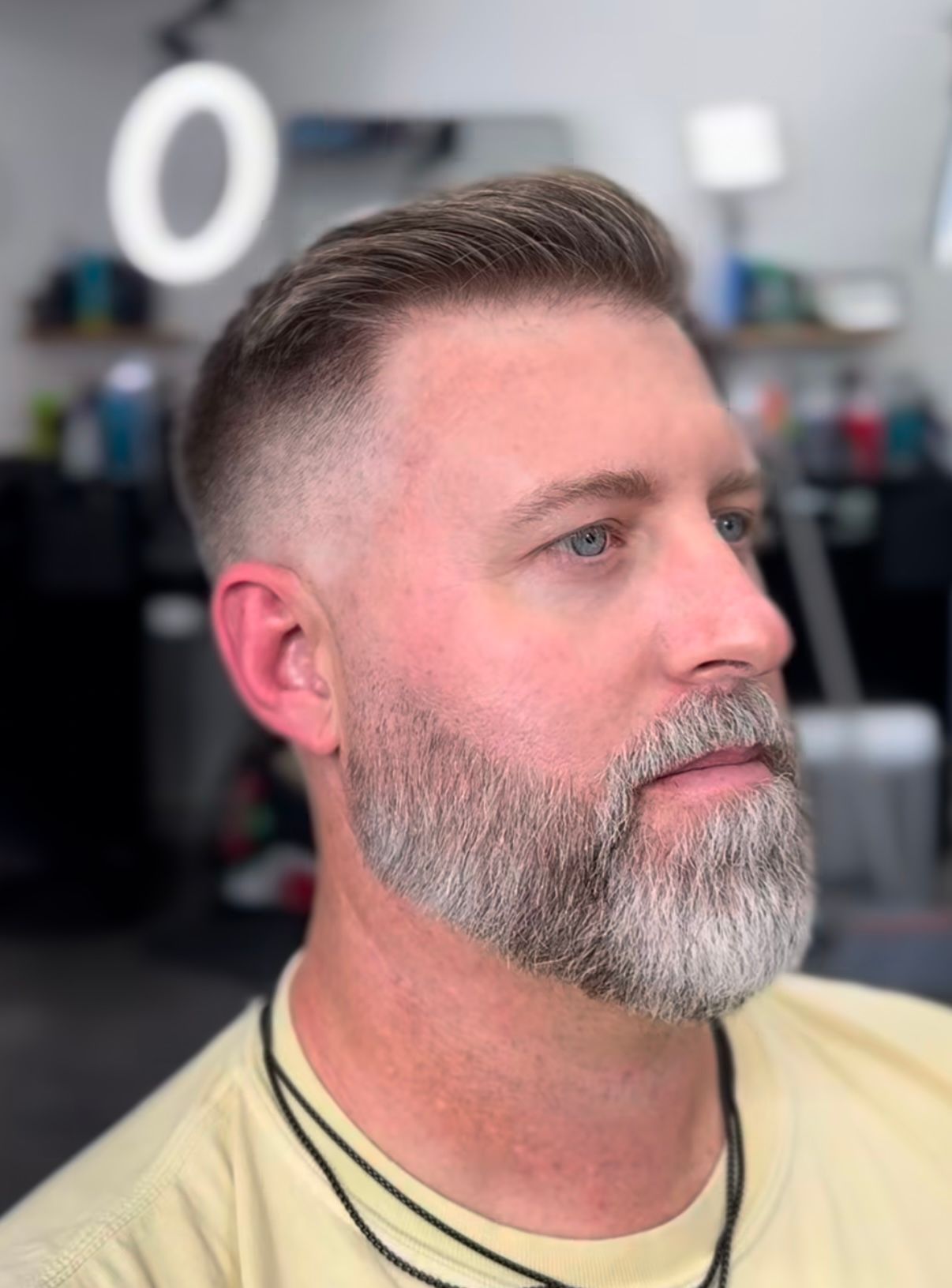 Man with short fade haircut and gray beard in a barbershop, looking to the side.