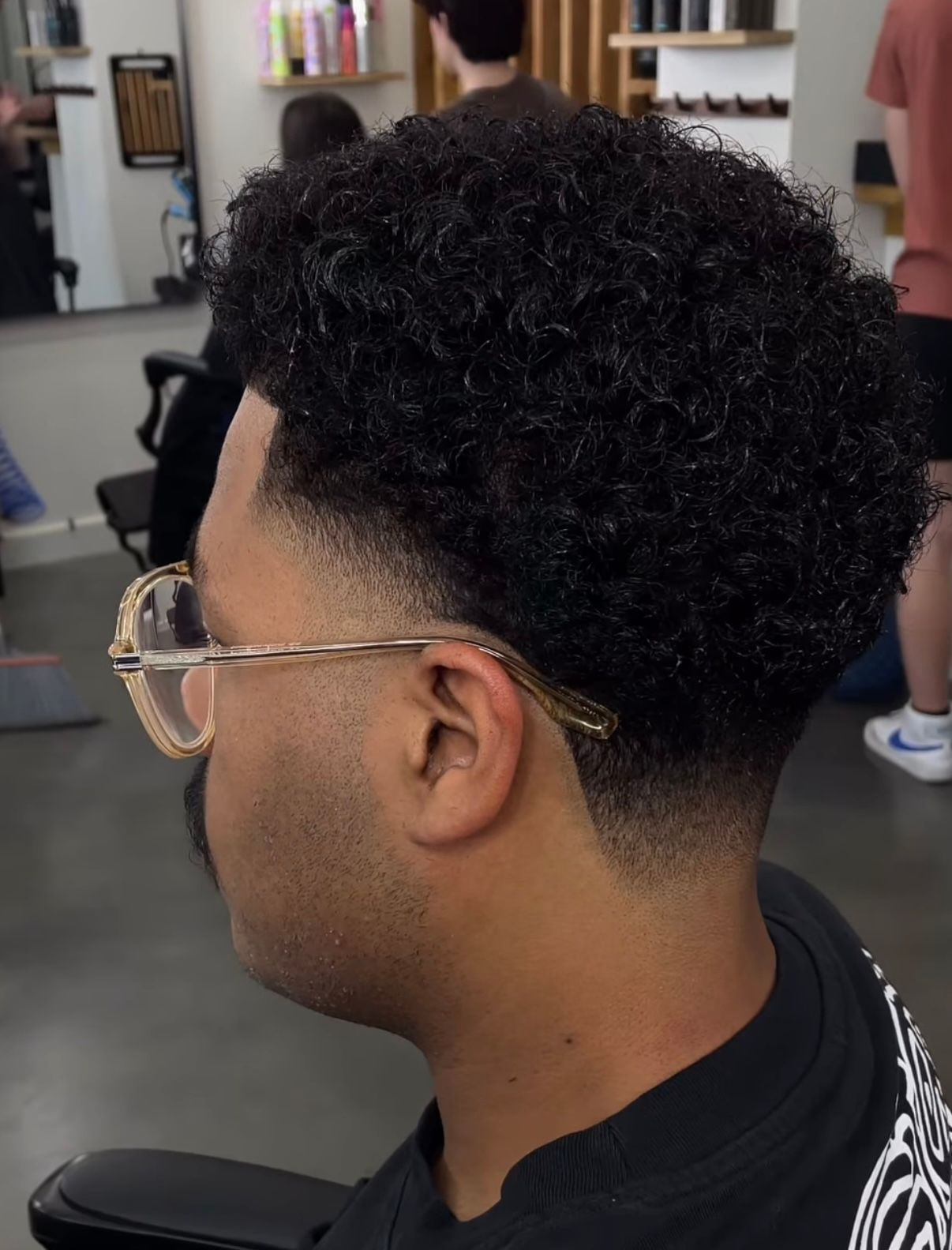 Side profile of a person with a short curly fade haircut and glasses in a barbershop.