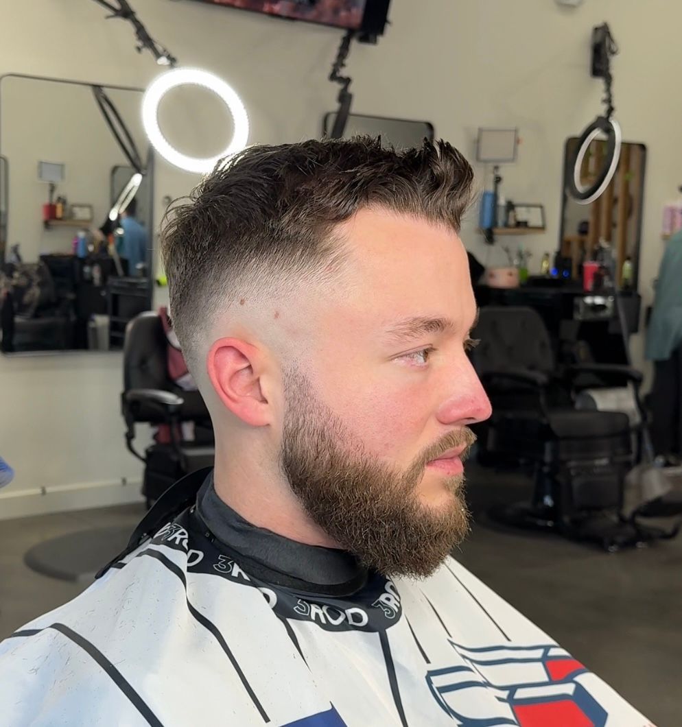 Profile view of a man with a short fade haircut and beard in a barbershop chair, with a ring light behind him
