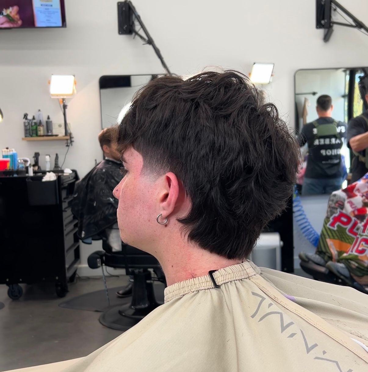 Short dark haircut with shaved sides in a salon chair, viewed from behind and side.