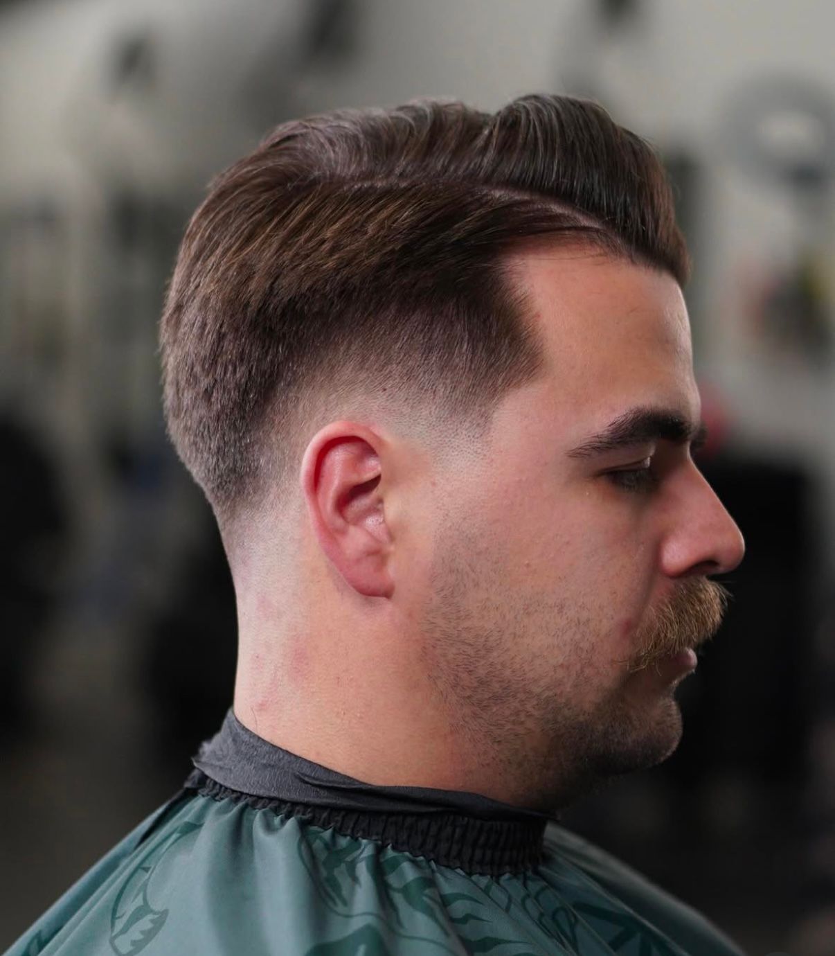 Side-profile of a man with a faded haircut and mustache, wearing a barber cape in a salon.