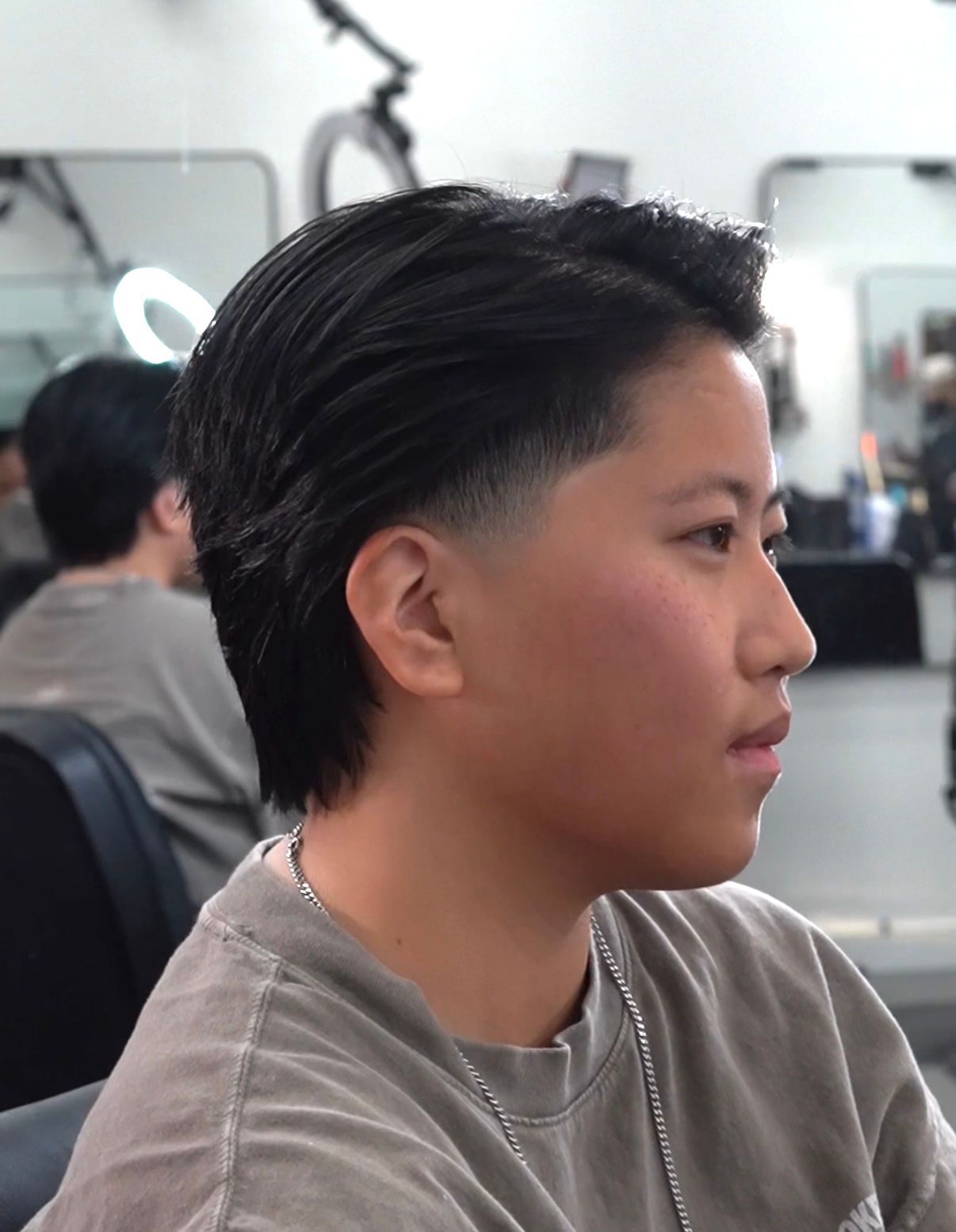 Profile view of a person with short dark hair in a gray shirt in a salon chair, with mirrors and styling tools behind them