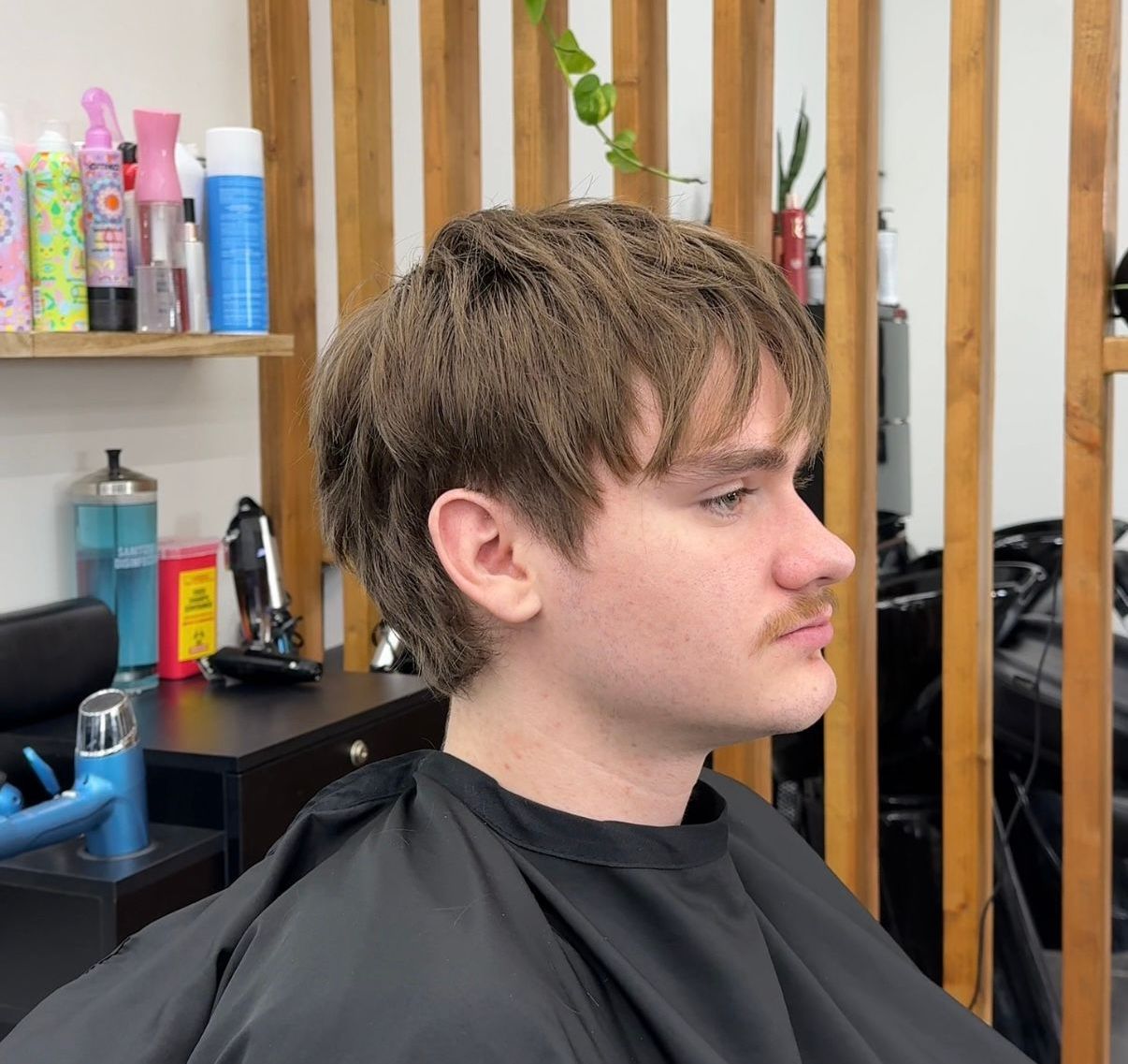 Young man with shaggy brown hair sitting in a barber chair, wearing a black salon cape