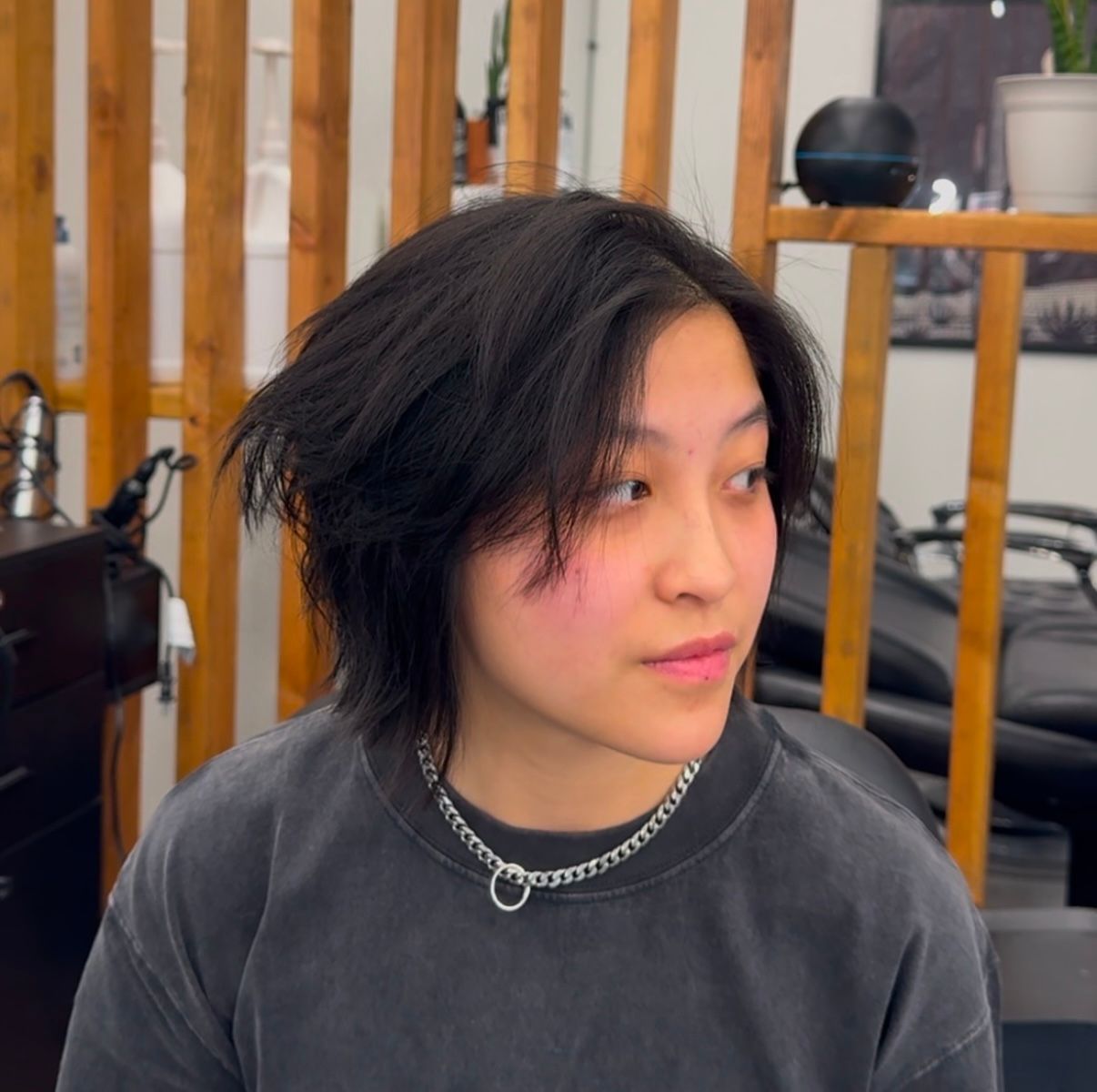 Person with short dark hair and silver chain necklace looks to the side in a studio setting.