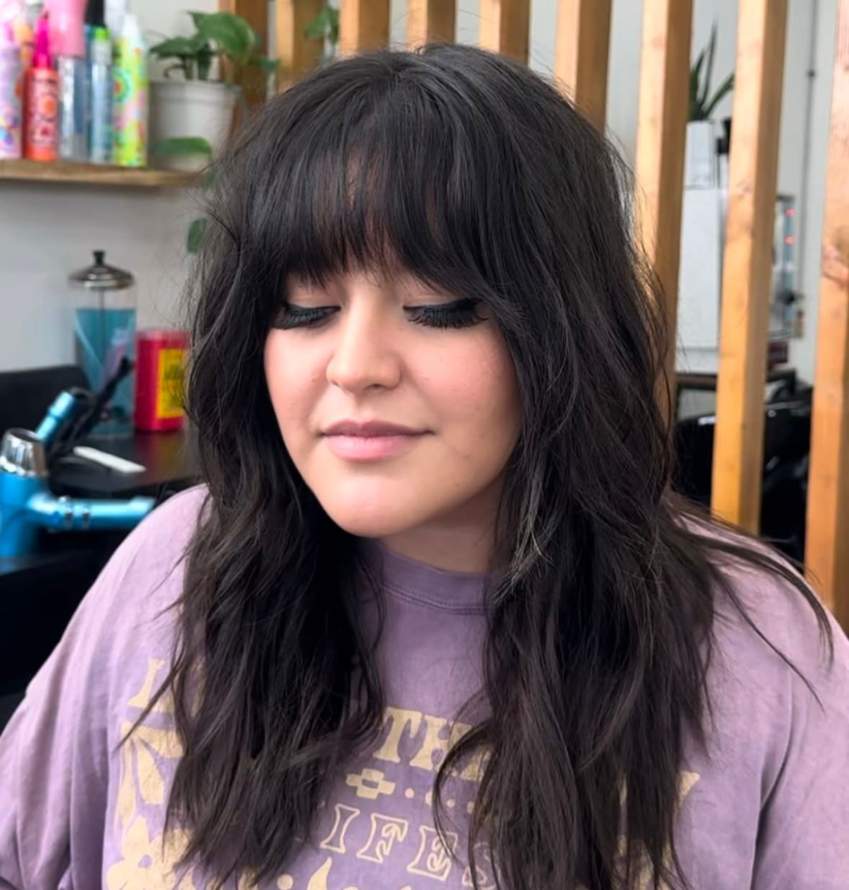 Close-up of a person with long dark hair and bangs wearing a lavender shirt, in a salon chair