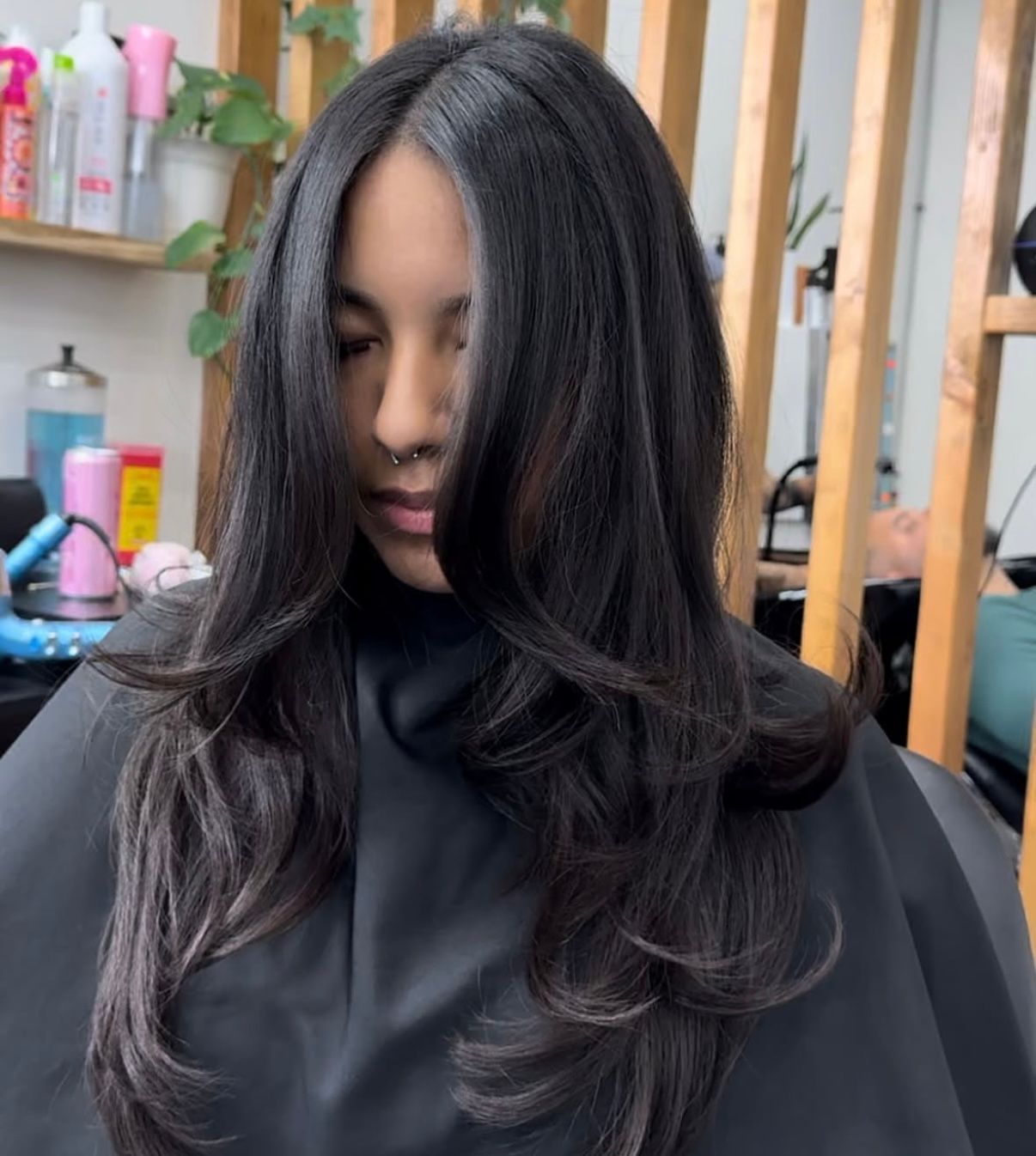 Person with long black hair in a salon chair, wearing a black cape, with layered ends and curtain bangs.