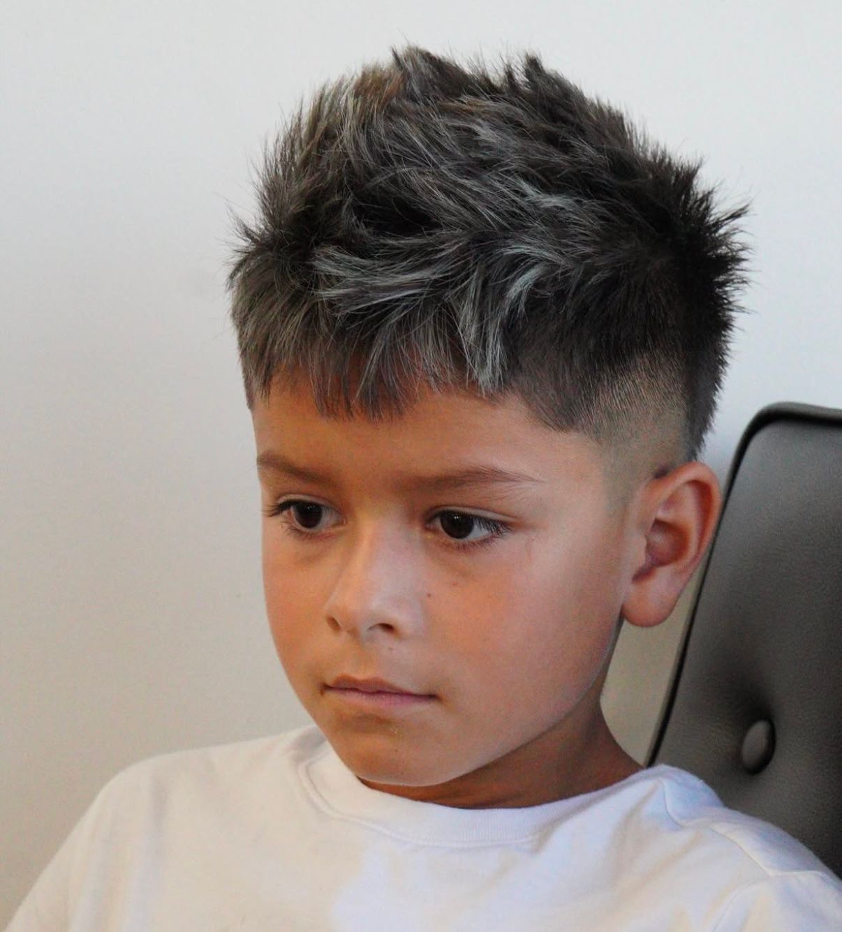 Young person with spiky dark hair wearing a white shirt, seated indoors with a neutral expression