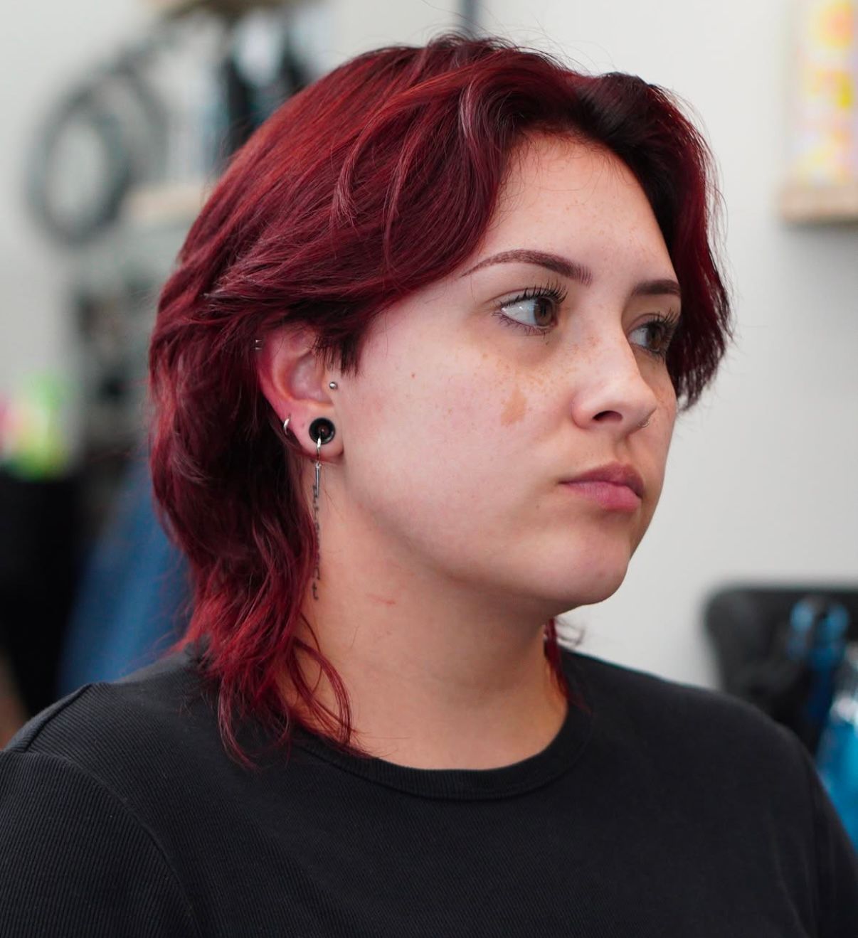 Person with short red hair and black shirt, shown in profile against a blurred indoor background