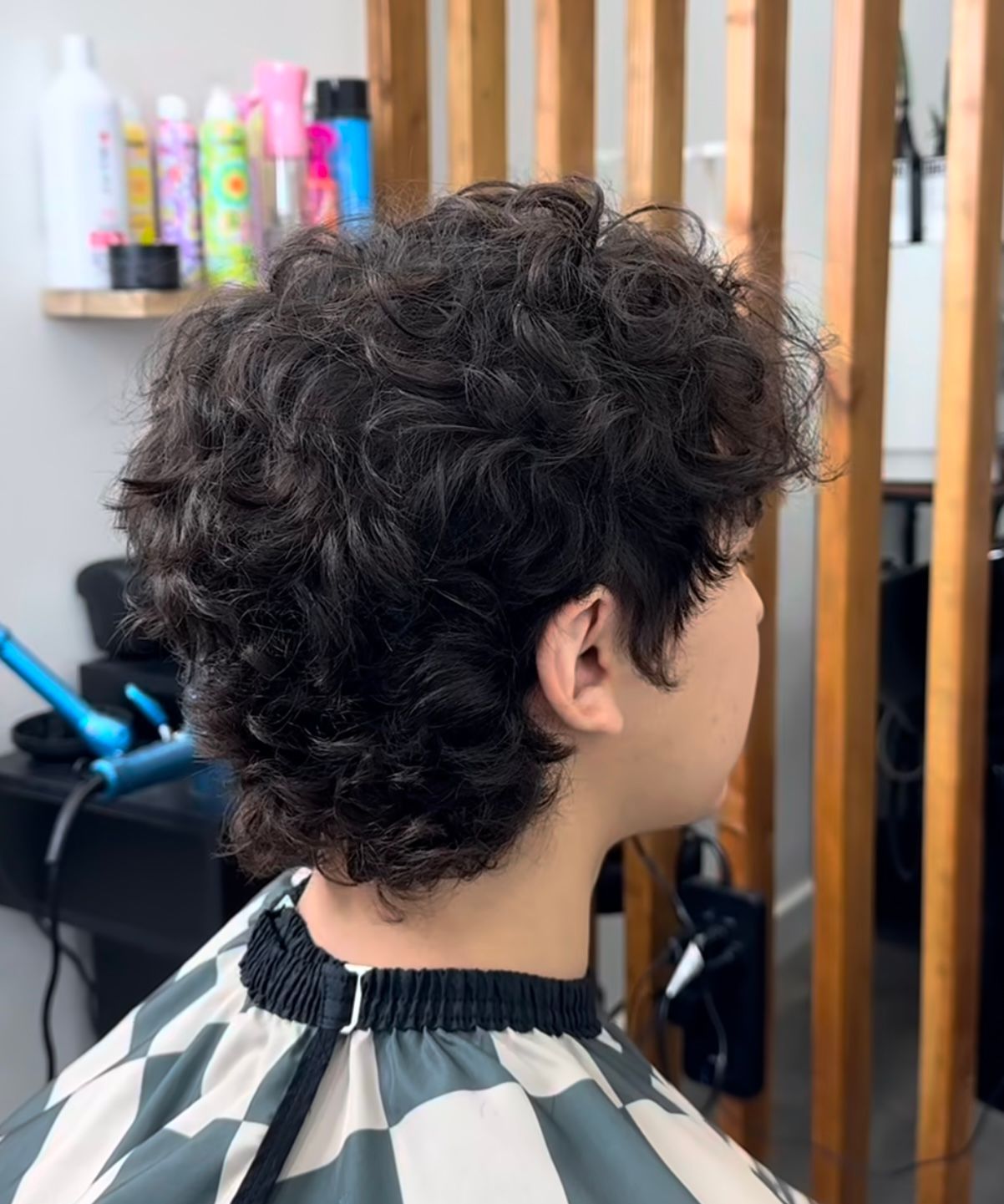 Curly-haired person in a salon chair, viewed from the side, with a striped cape around the neck.