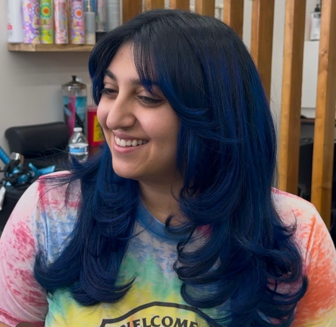 Smiling person with blue hair in a colorful shirt, standing in a salon-like setting