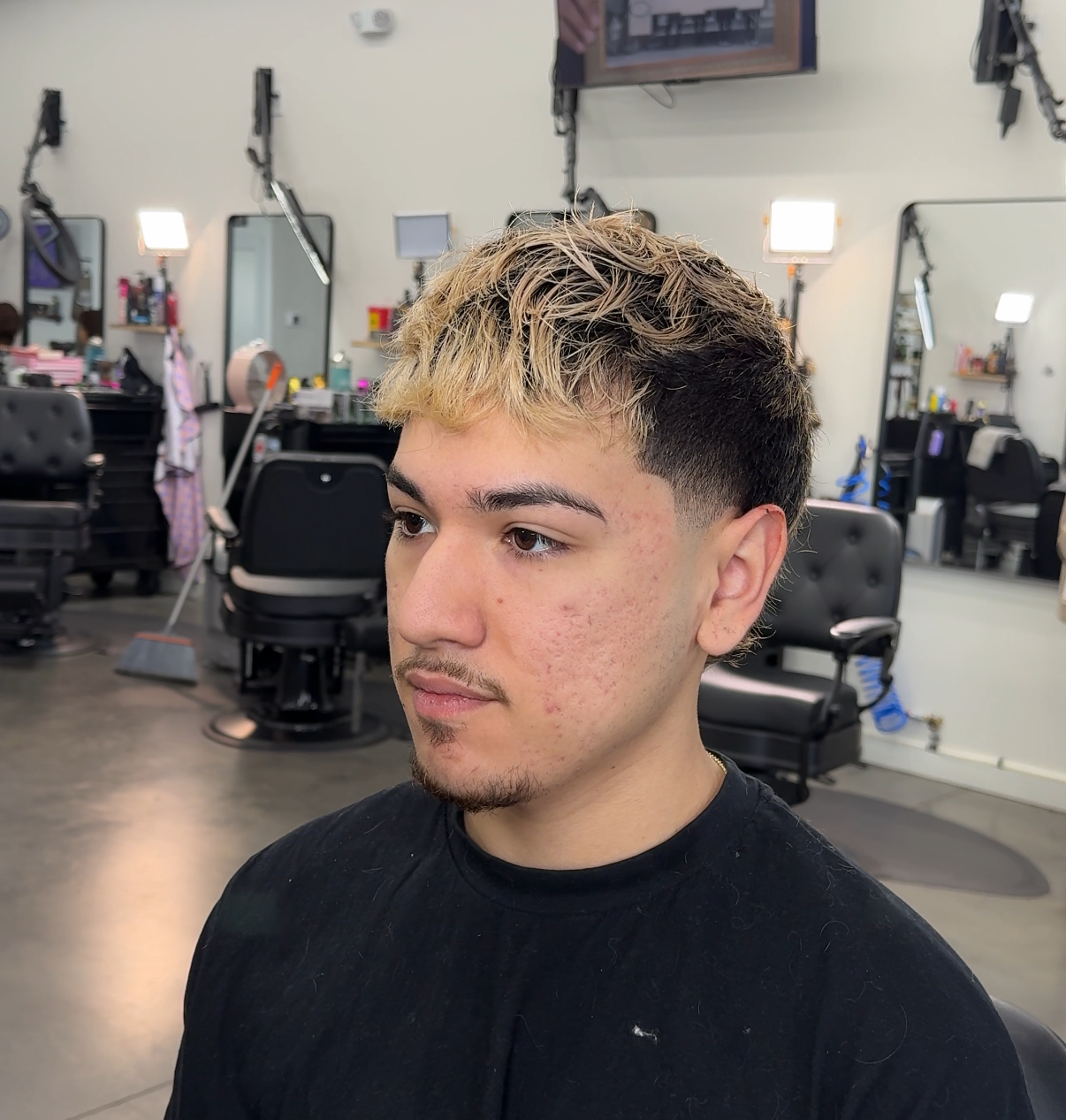 Person with curly blond hair in a barber shop, wearing a black shirt, looking to the side.