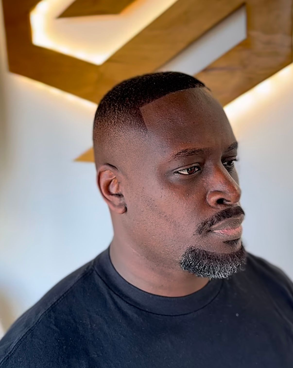 Man in a dark shirt with a short fade haircut, looking to the side indoors under warm lighting