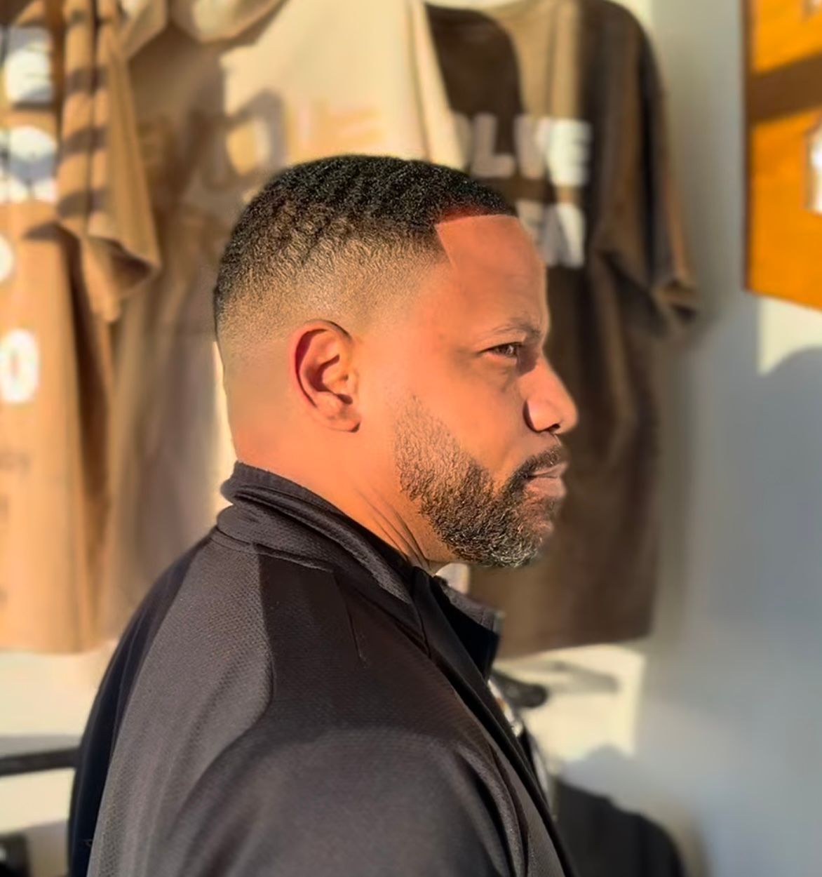 Side-profile portrait of a man in a dark jacket, standing in warm sunlight near hanging T-shirts.