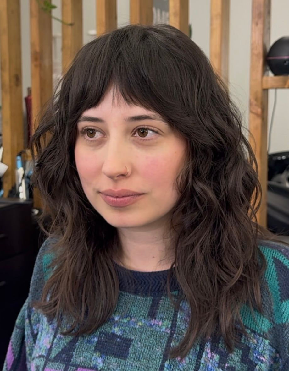 Portrait of a woman with wavy dark hair and bangs, wearing a patterned sweater indoors