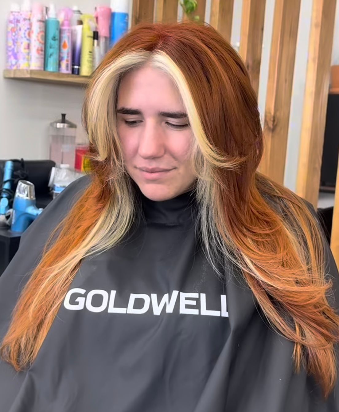 Woman with long red-and-blonde hair sitting in a salon chair, wearing a Goldwell cape and smiling softly.