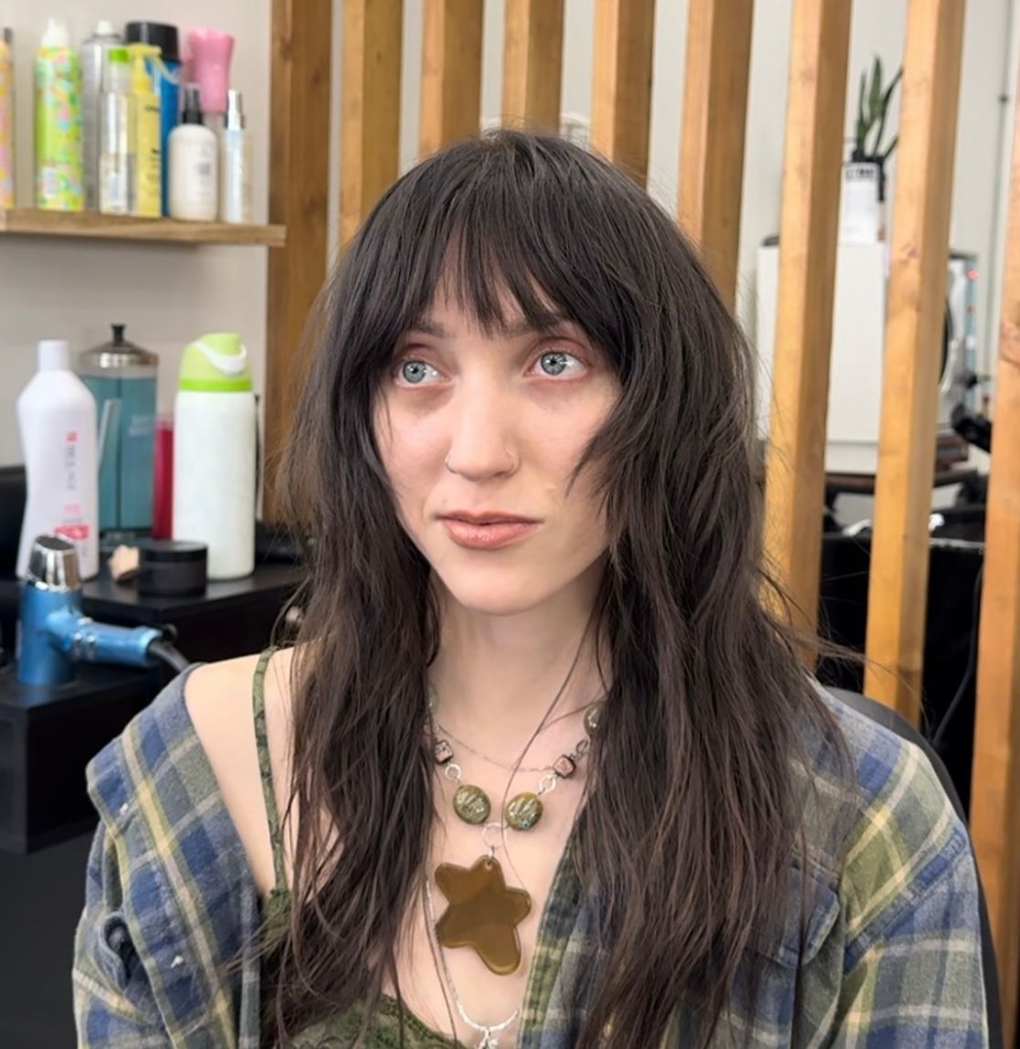 Woman with dark hair sitting in a salon, wearing a plaid top and pendant necklace.
