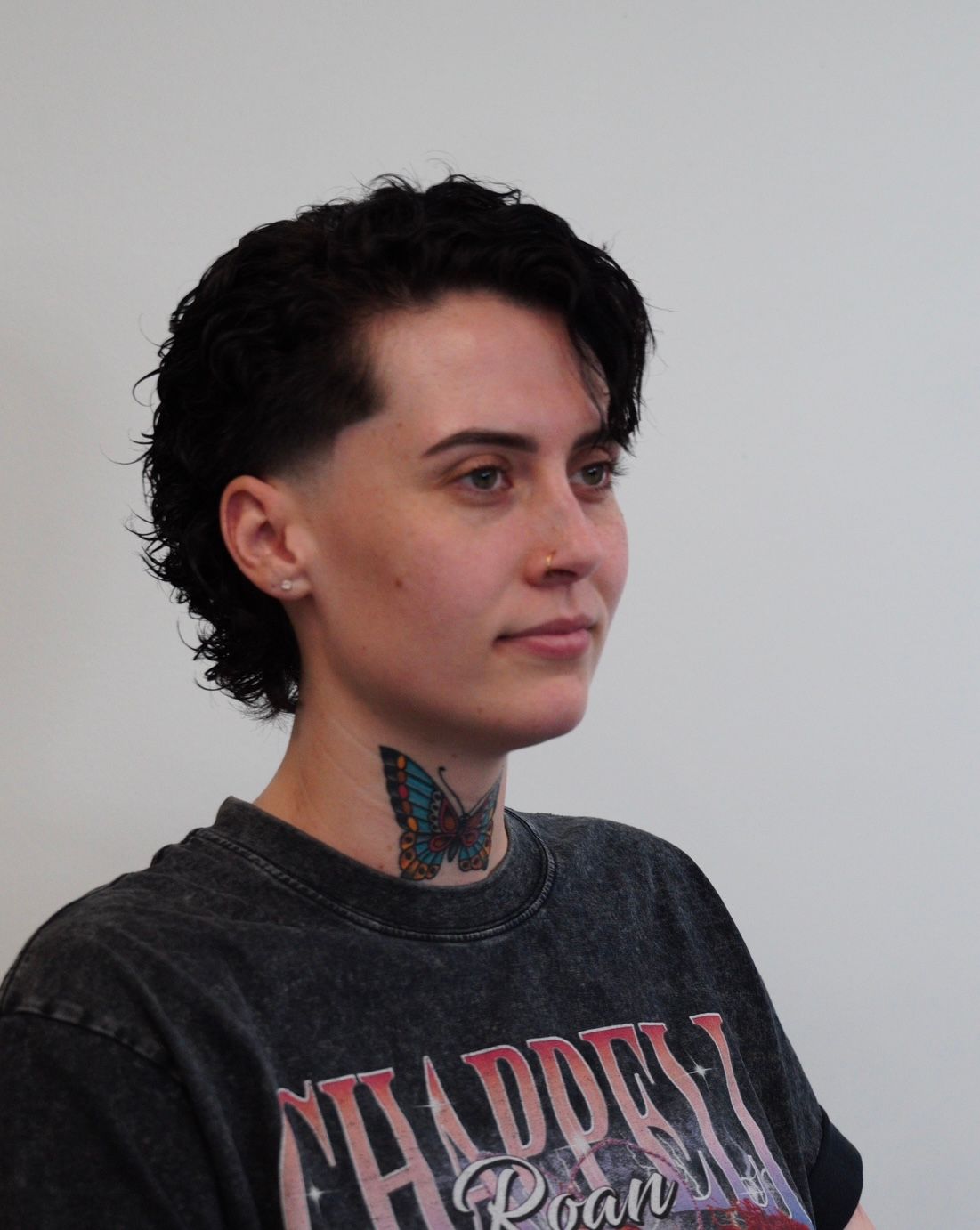 Person with short dark hair and neck tattoo wearing a dark graphic T-shirt, in profile against a plain wall