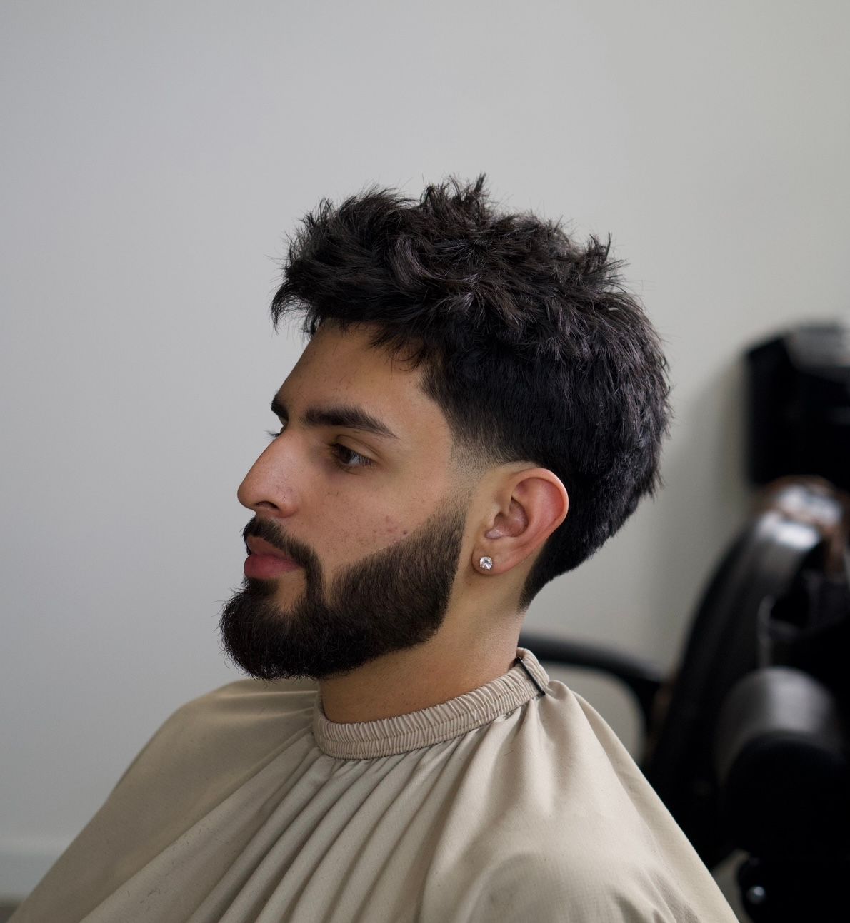 Profile view of a bearded man with short curly hair and a beige shirt, seated indoors on a neutral background