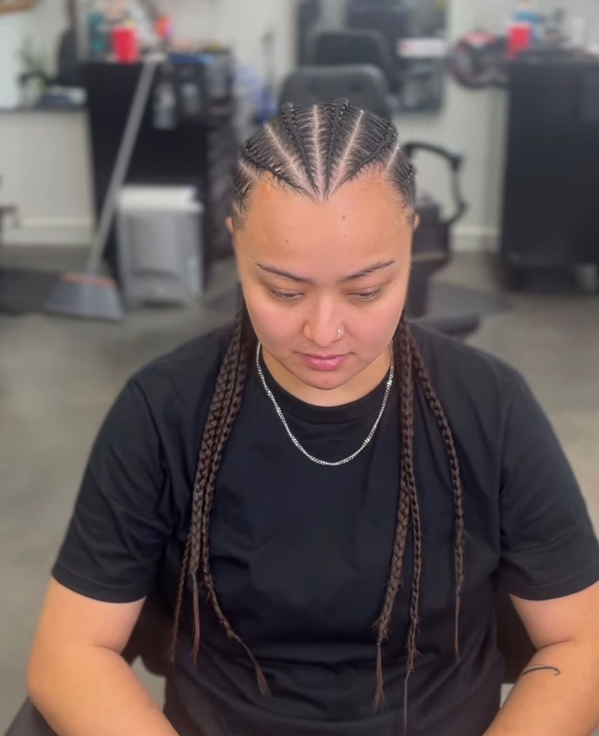 Person with braided hair looking down, wearing a black shirt in a salon chair.