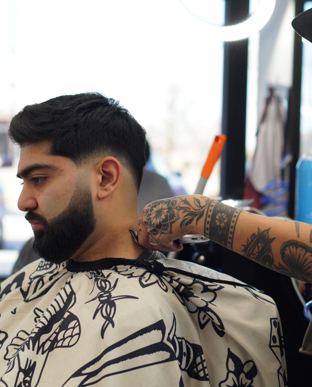 Man getting a beard trim in a barber chair, wearing a patterned cape, with a tattooed arm in frame