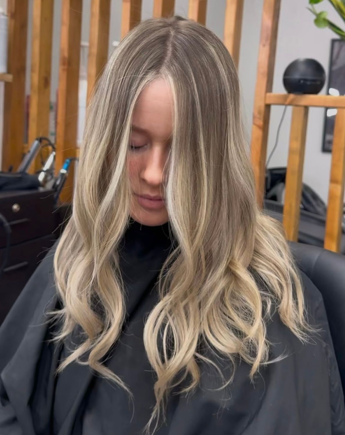 Person with long wavy blonde hair and dark roots sitting in a salon chair, eyes closed.