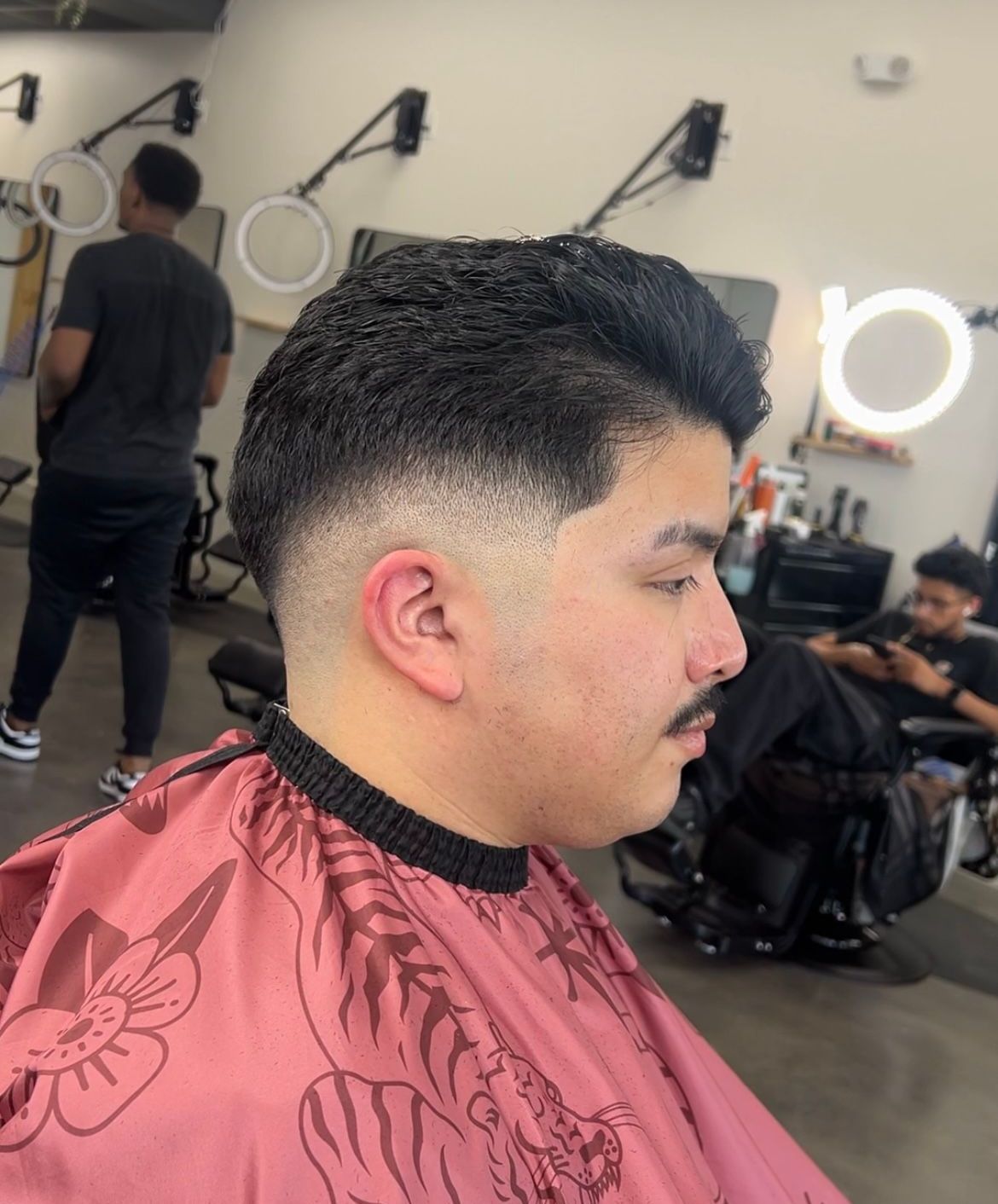 Profile view of a man getting a fresh fade haircut in a barbershop, wearing a pink cape