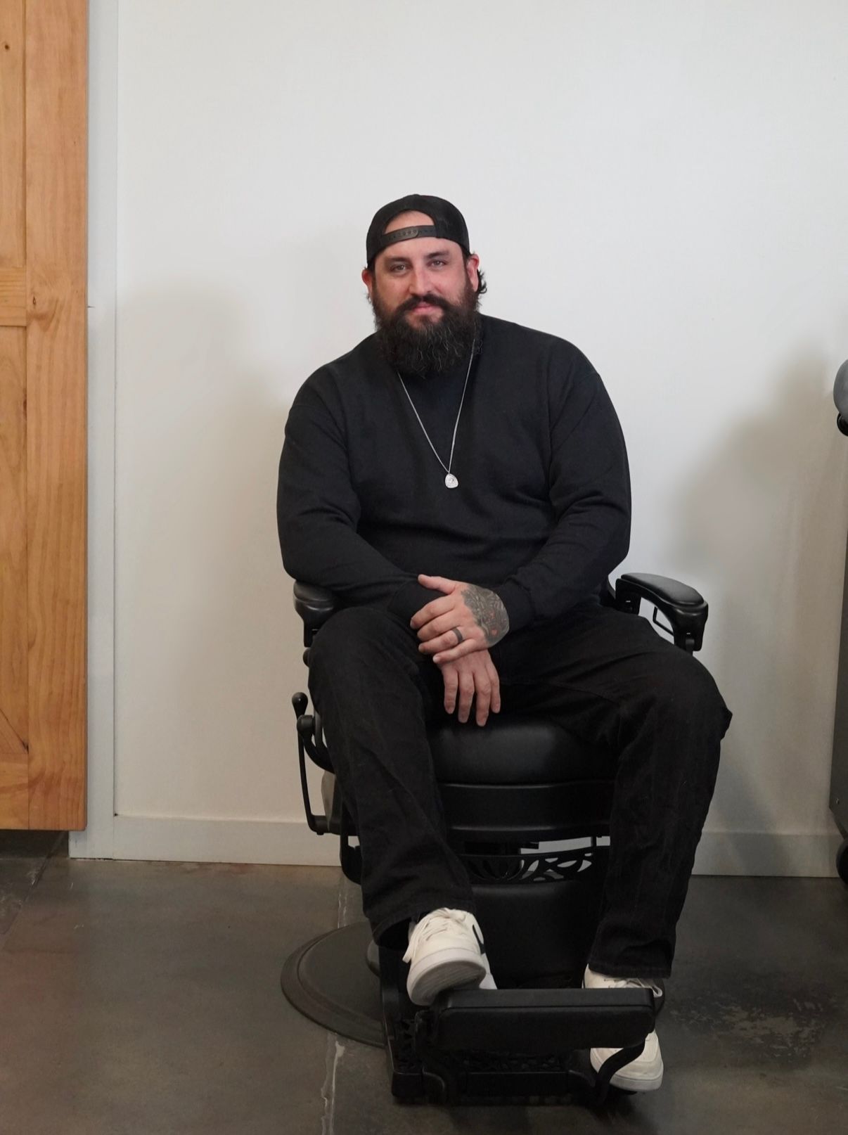 Man seated in a black chair against a plain wall, wearing black clothes and a backward cap.