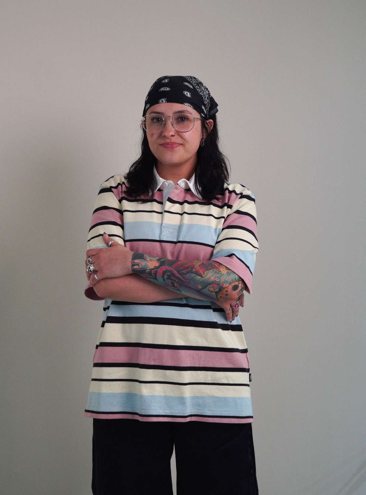 Person standing with arms crossed, wearing a striped shirt, black bandana, and glasses against a plain wall.