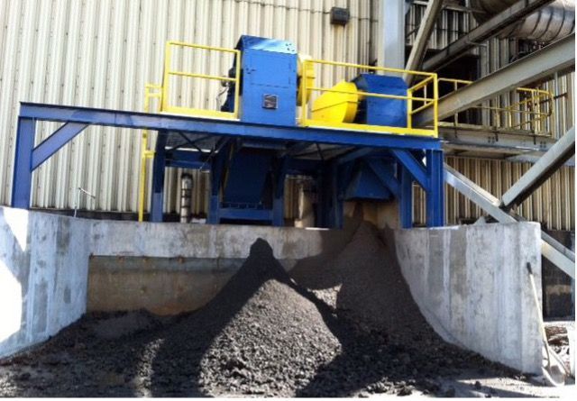 Blue and yellow industrial machinery atop a blue frame, dispensing dark material onto a pile in a concrete structure.