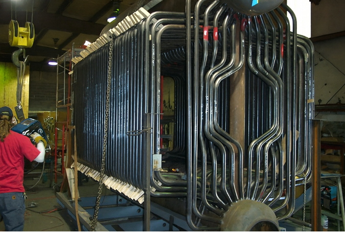 Worker welding metal tubes in a factory, likely constructing a heat exchanger. A large, black-tubed structure is visible.