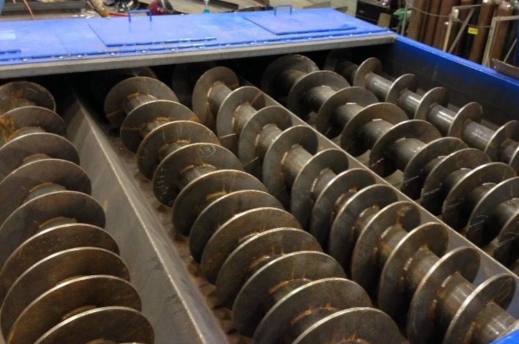 Close-up of industrial equipment: multiple rotating augers in a blue-colored trough, likely for material conveyance.