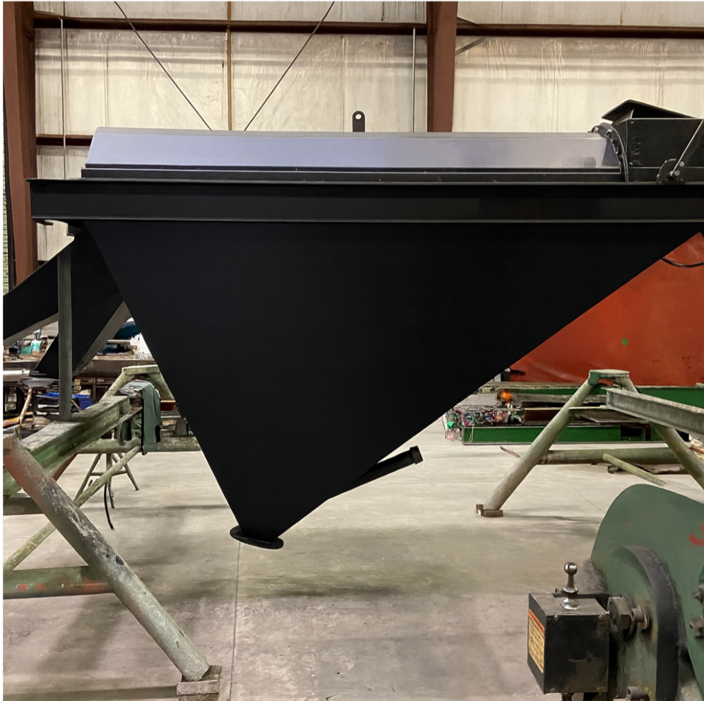 Black industrial hopper suspended indoors, with metal supports and a discharge tube.
