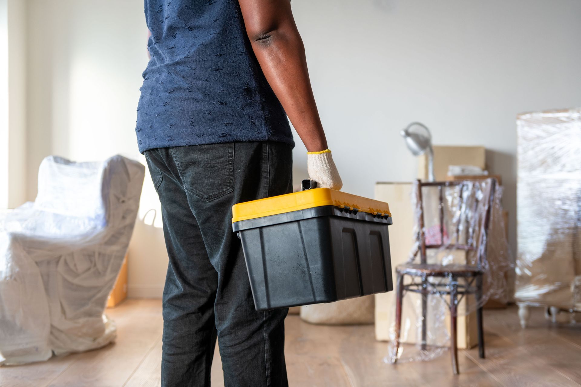 A cropped view of a man holding a toolbox in a cluttered room of wrapped furniture & cardboard boxes