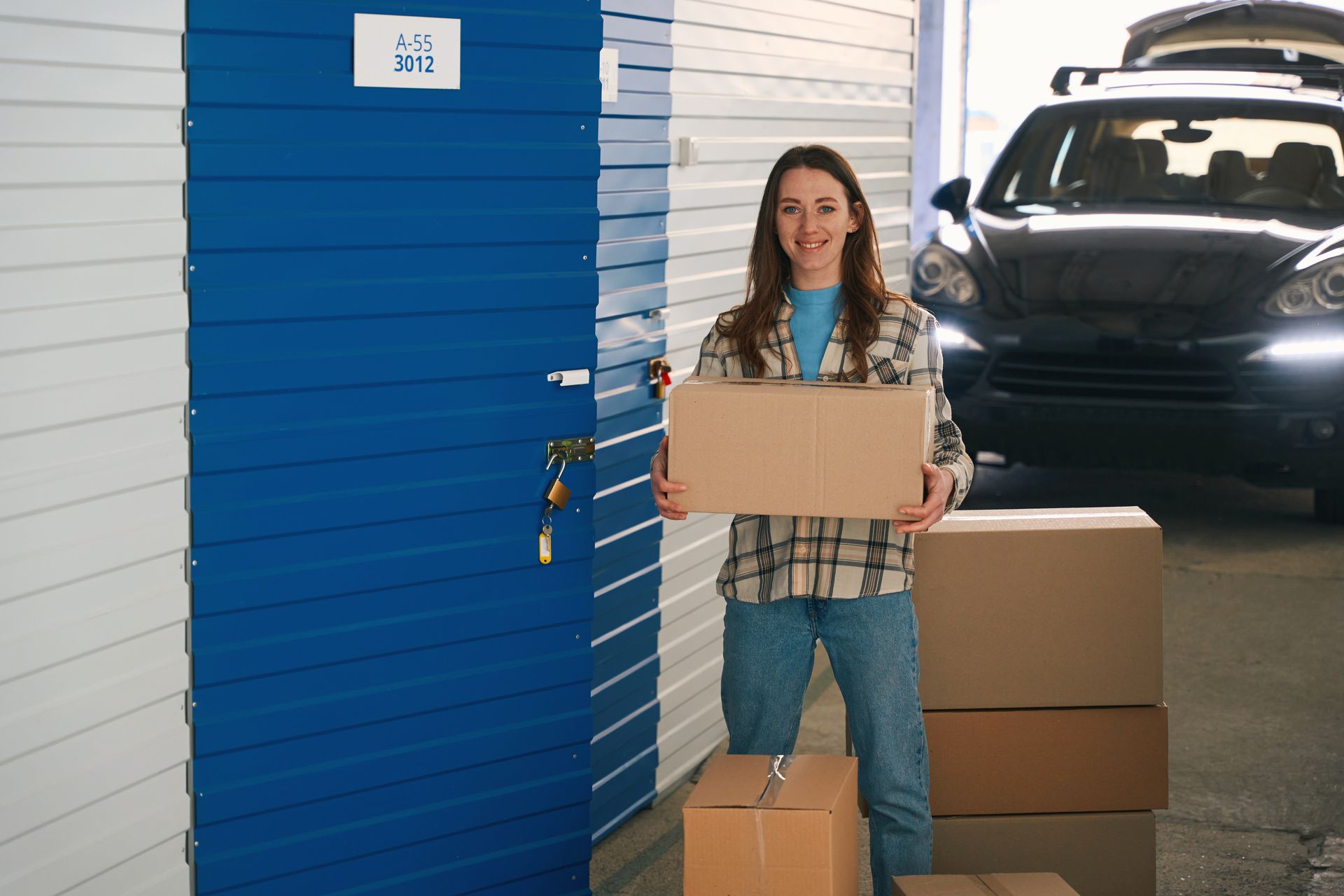 A woman with big cardboard boxes next to an open self-storage unit. A black car is in the background.