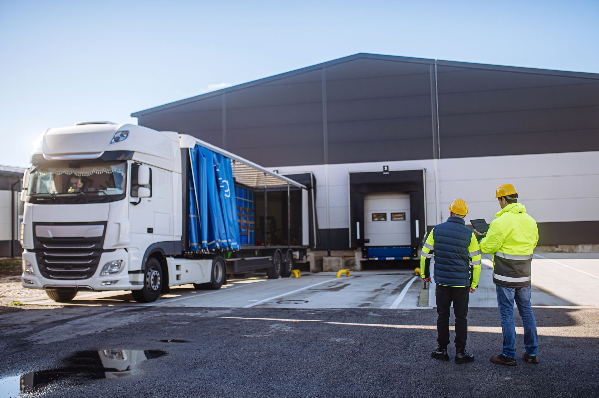 Two workers in safety gear observe a white semi-truck unloading goods at a warehouse loading dock.