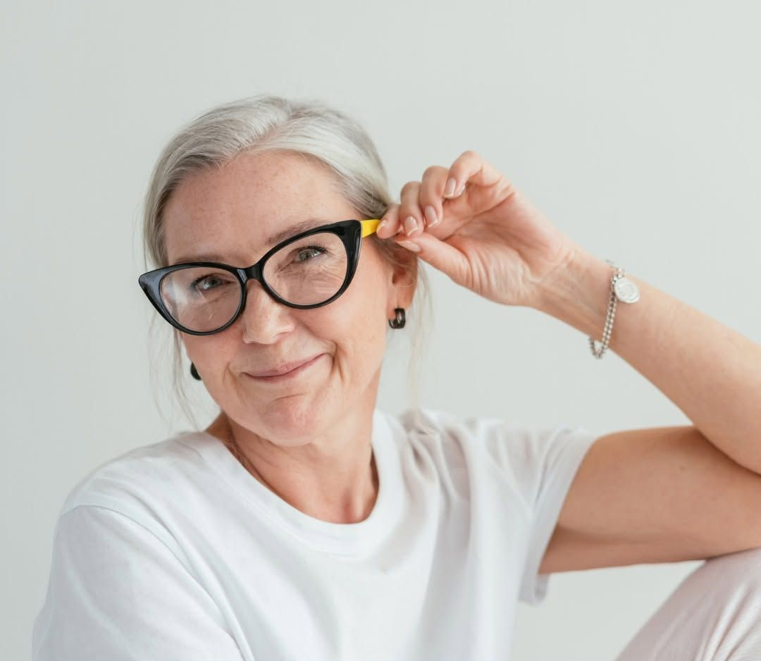 Persona sorridente che si sistema gli occhiali neri, con indosso una camicia bianca, su sfondo chiaro.