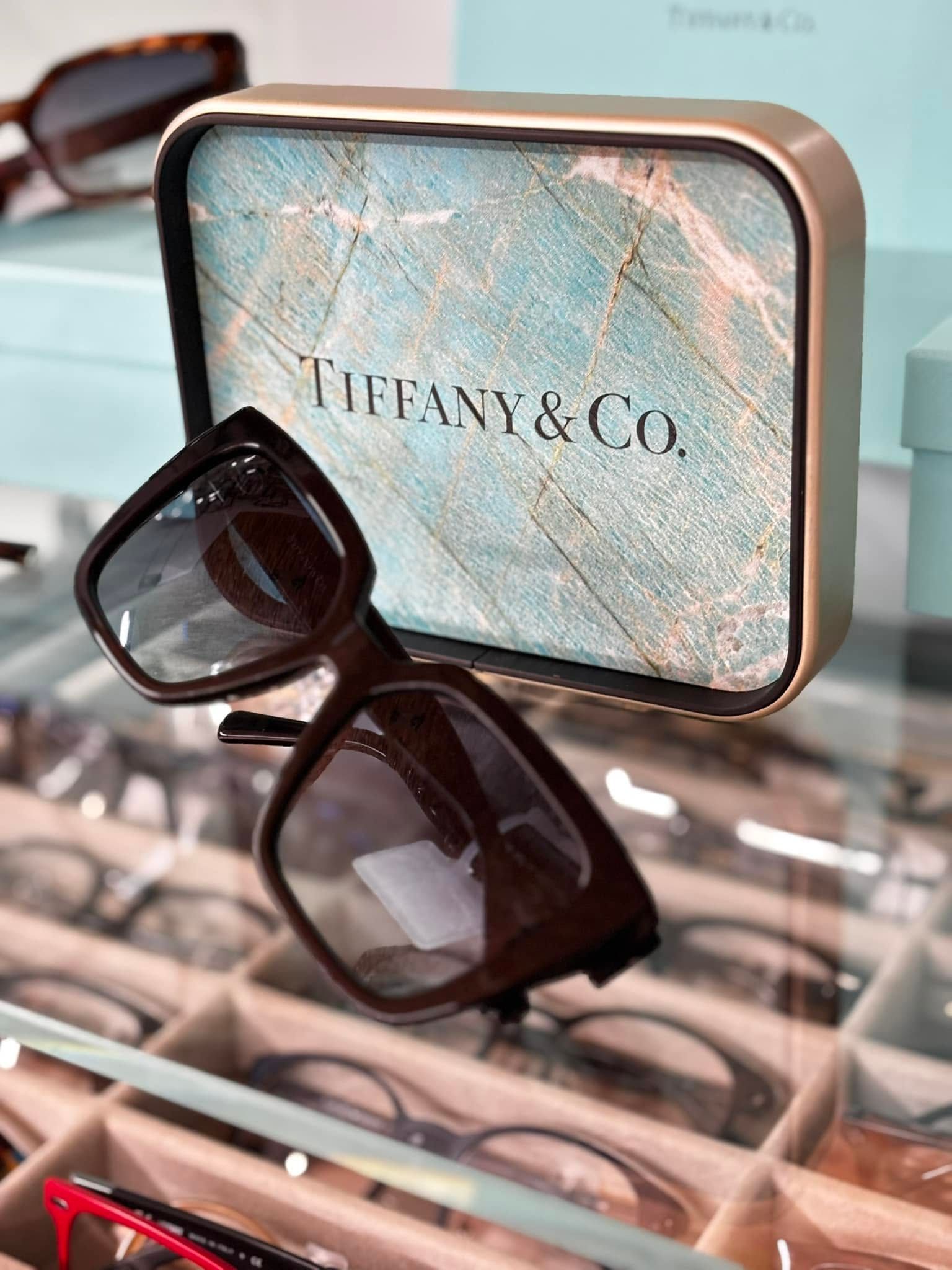 Occhiali da vista neri appoggiati su una custodia Tiffany & Co. sopra un vassoio espositivo con altri occhiali.