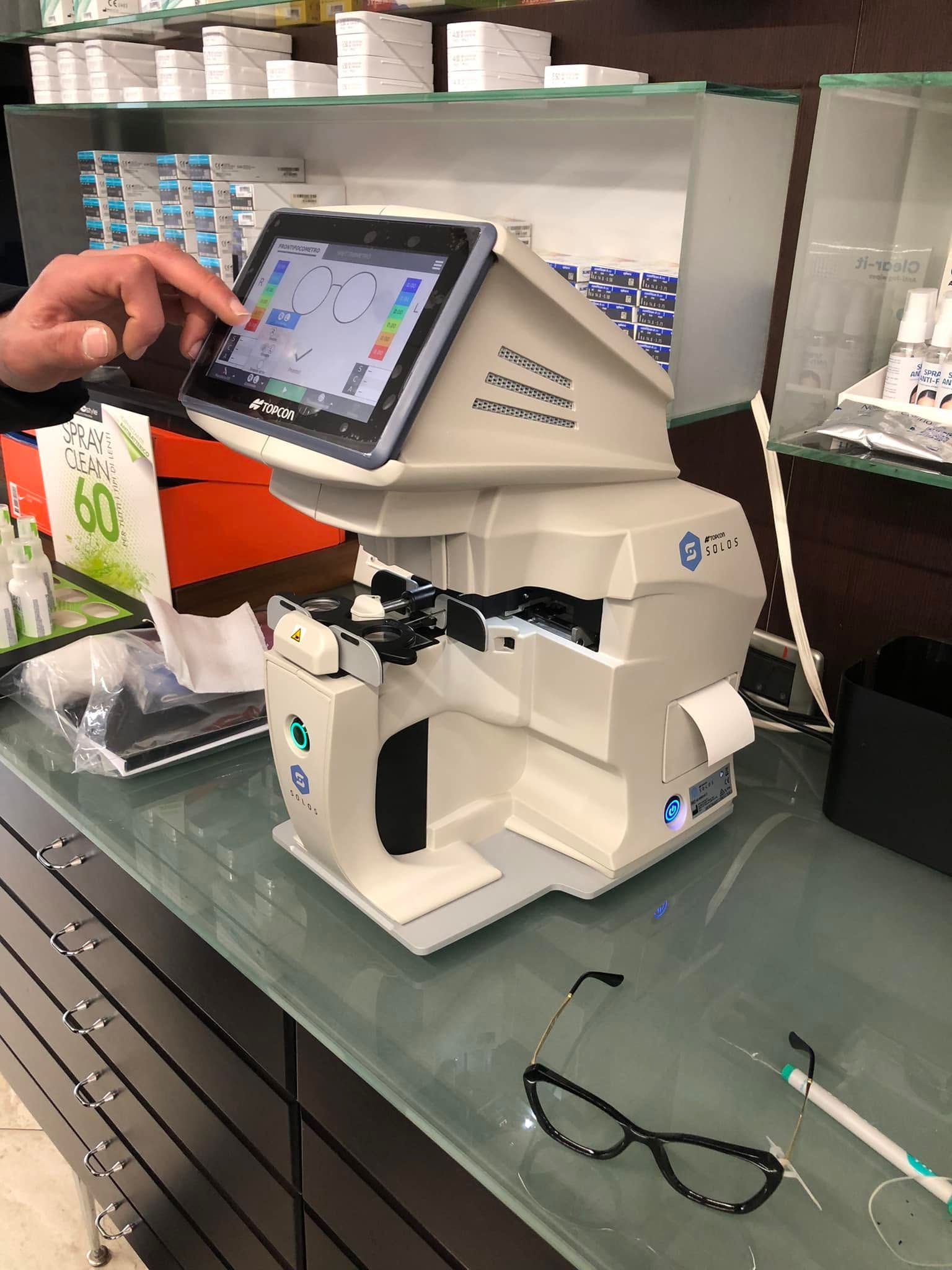 Una persona utilizza un misuratore di lenti a contatto con schermo tattile sul bancone di una farmacia, con degli occhiali nelle vicinanze.