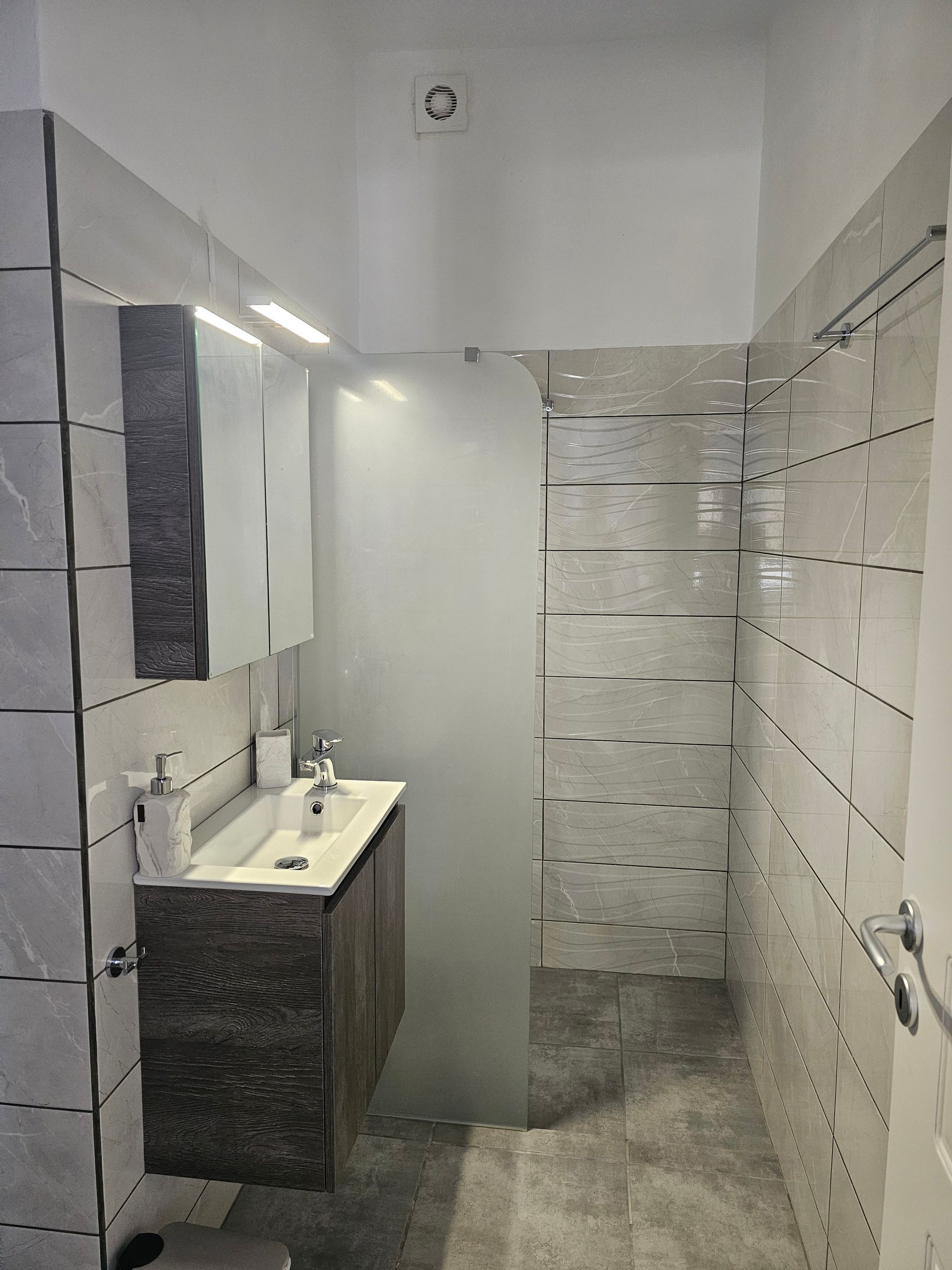 Narrow tiled bathroom with sink vanity, mirror cabinet, and shower area with frosted glass door.