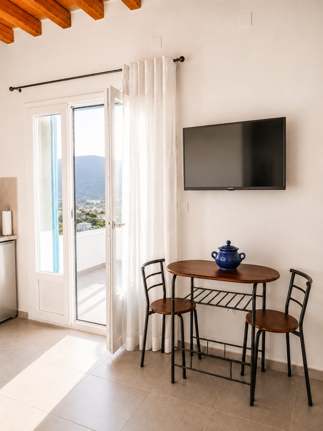 Small dining nook with round table, two chairs, wall-mounted TV, and open doors to a balcony with sheer curtains.