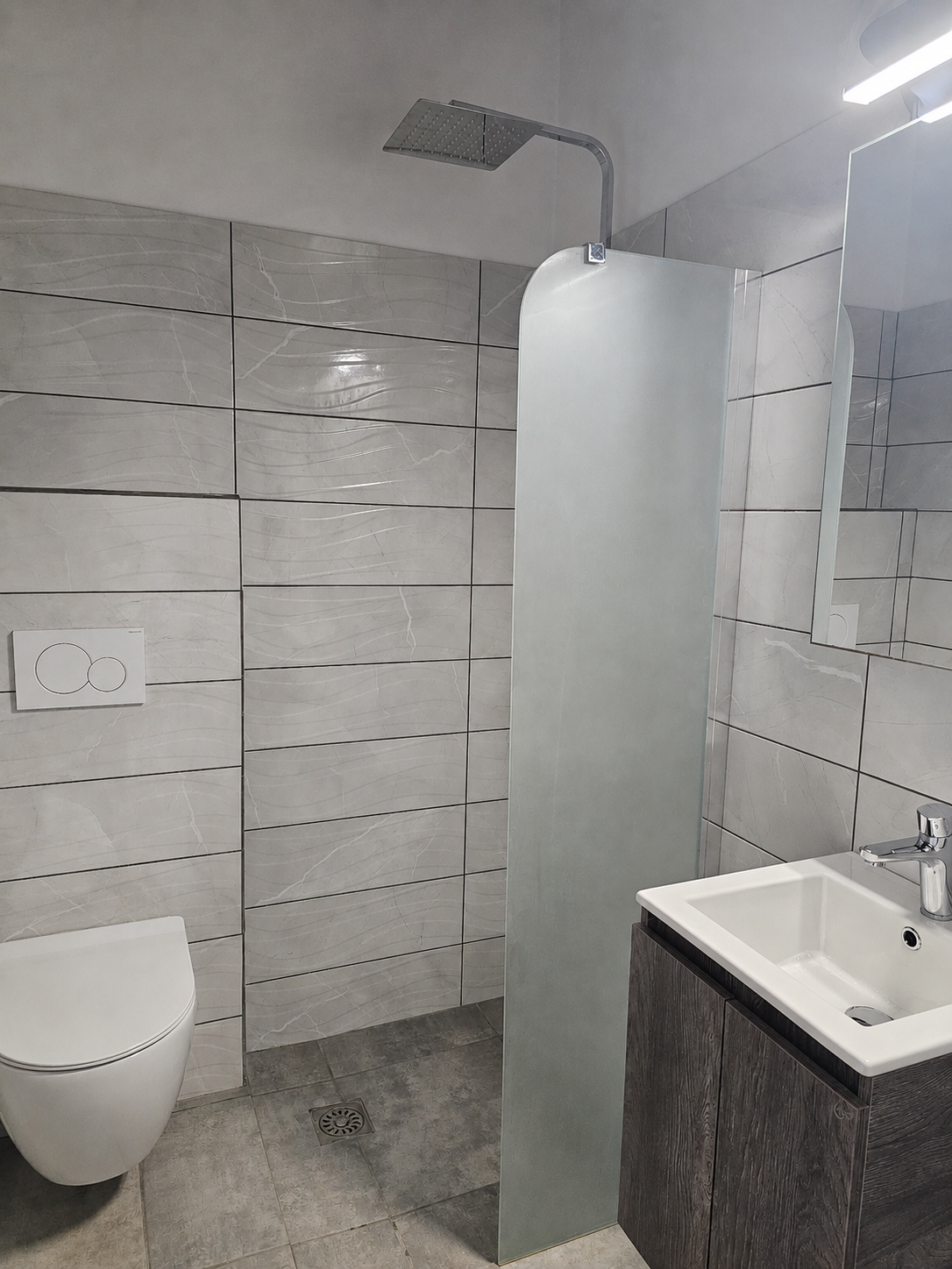 Small tiled bathroom with wall-mounted toilet, frosted shower partition, and white sink.