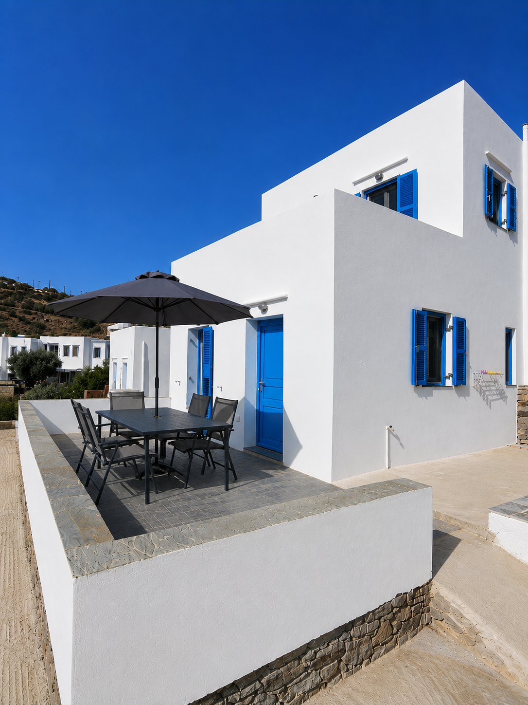 White modern house with blue shutters and patio furniture under a black umbrella by the sea