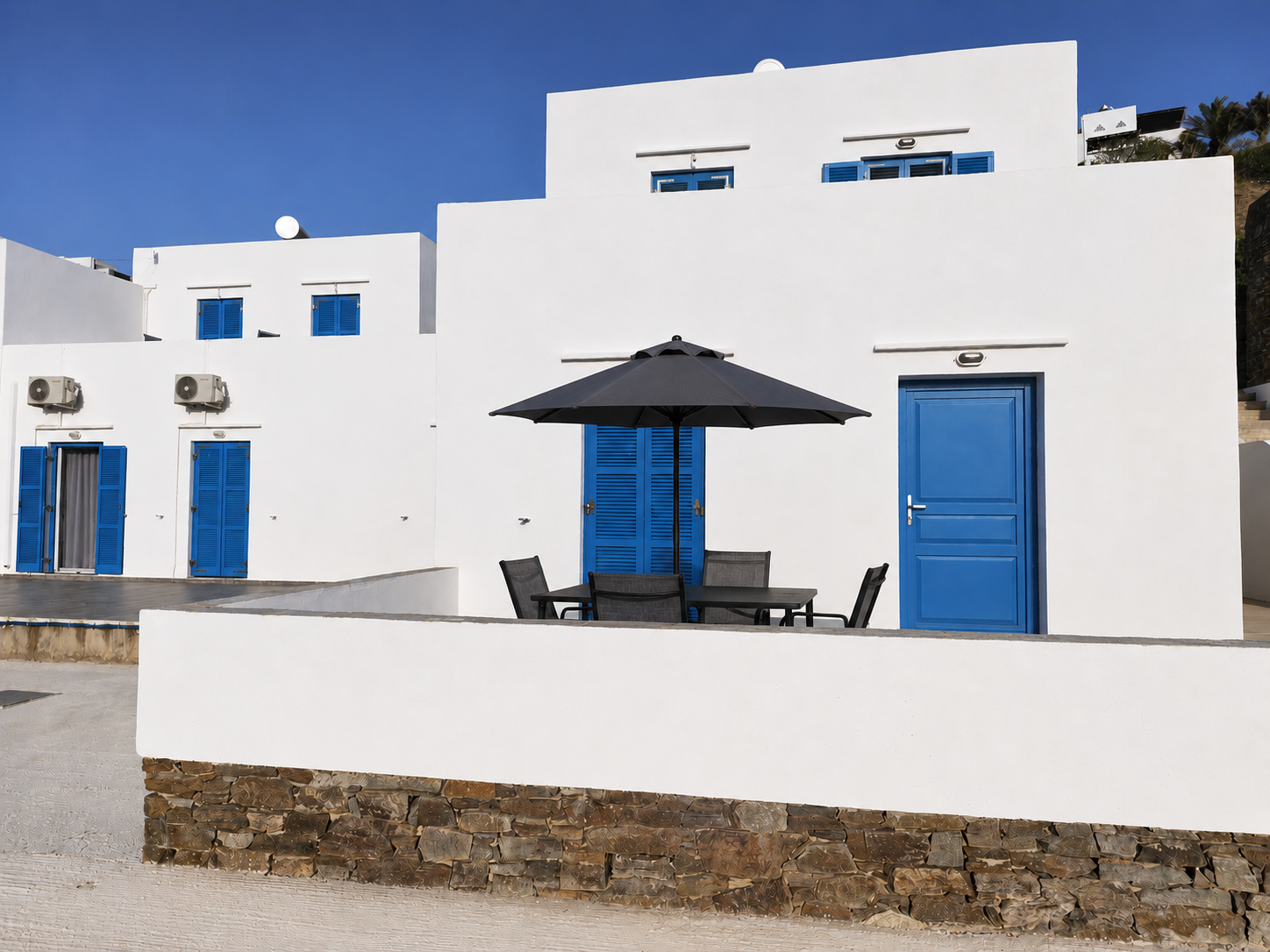 White seaside building with blue doors and windows, patio table under black umbrella, stone wall in front
