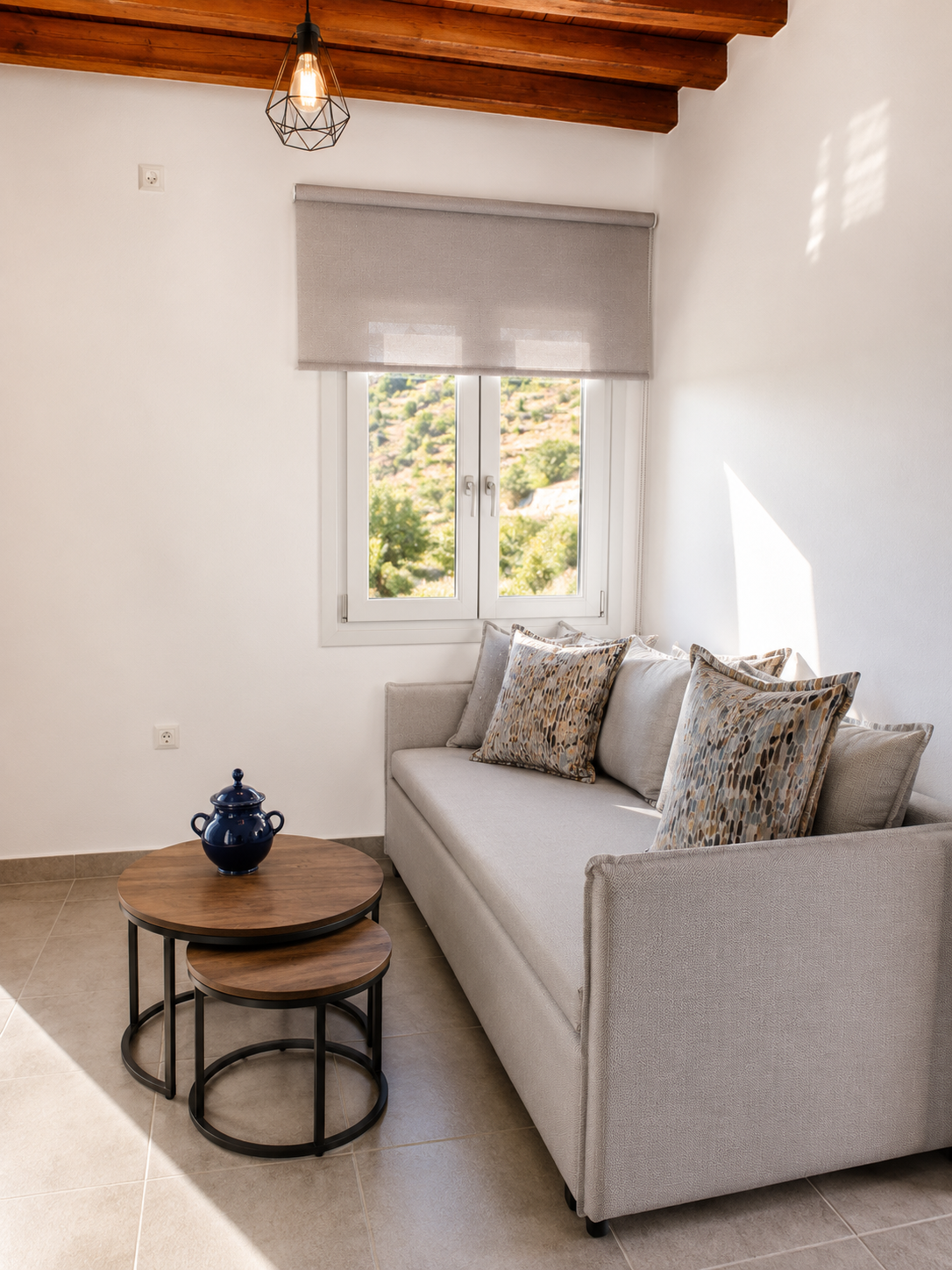 Small bright room with a gray sofa, round wooden tables, and a window with a blind.
