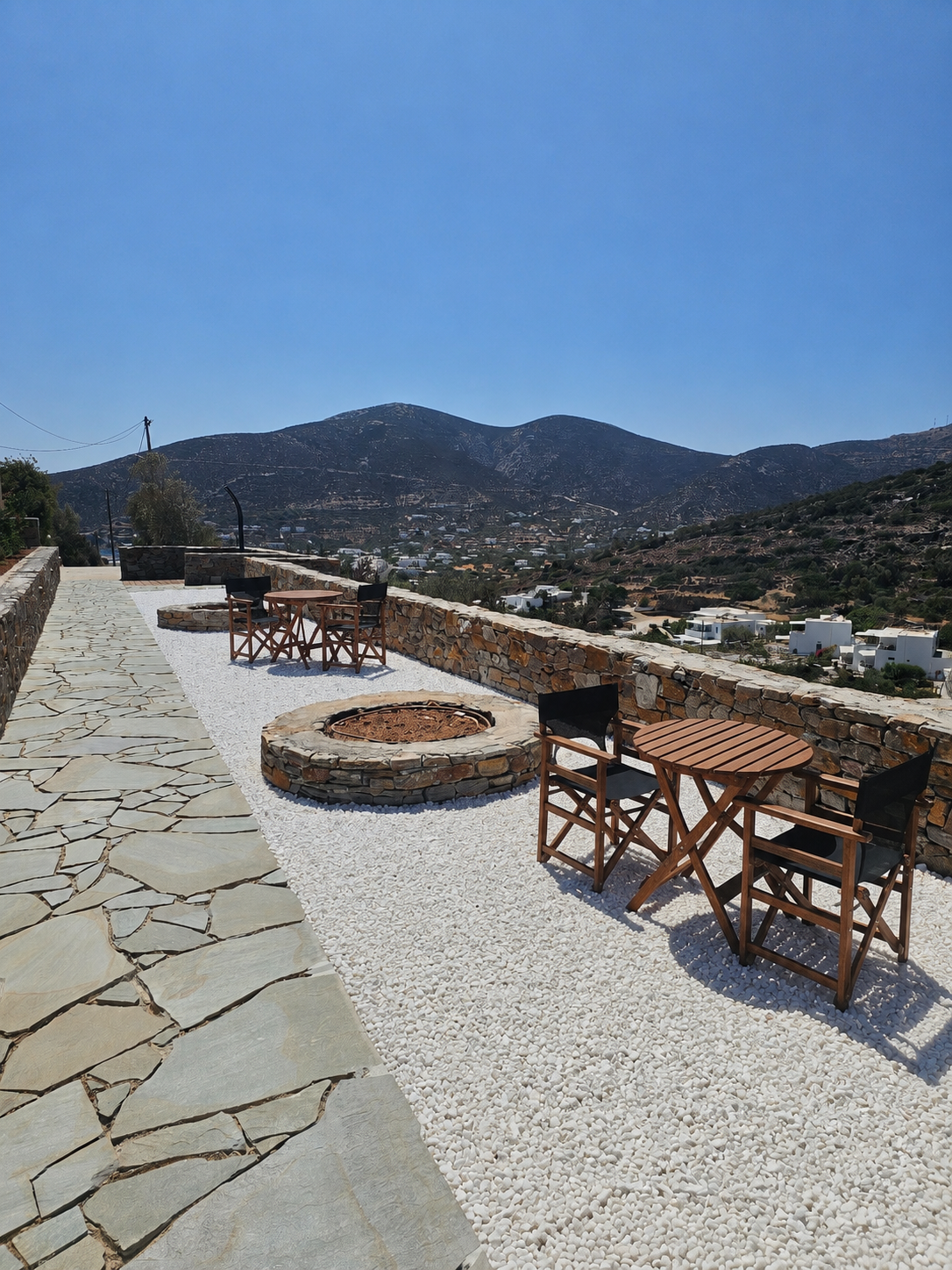 Outdoor terrace with tables, circular fire pit, stone path, and mountain view under a clear blue sky