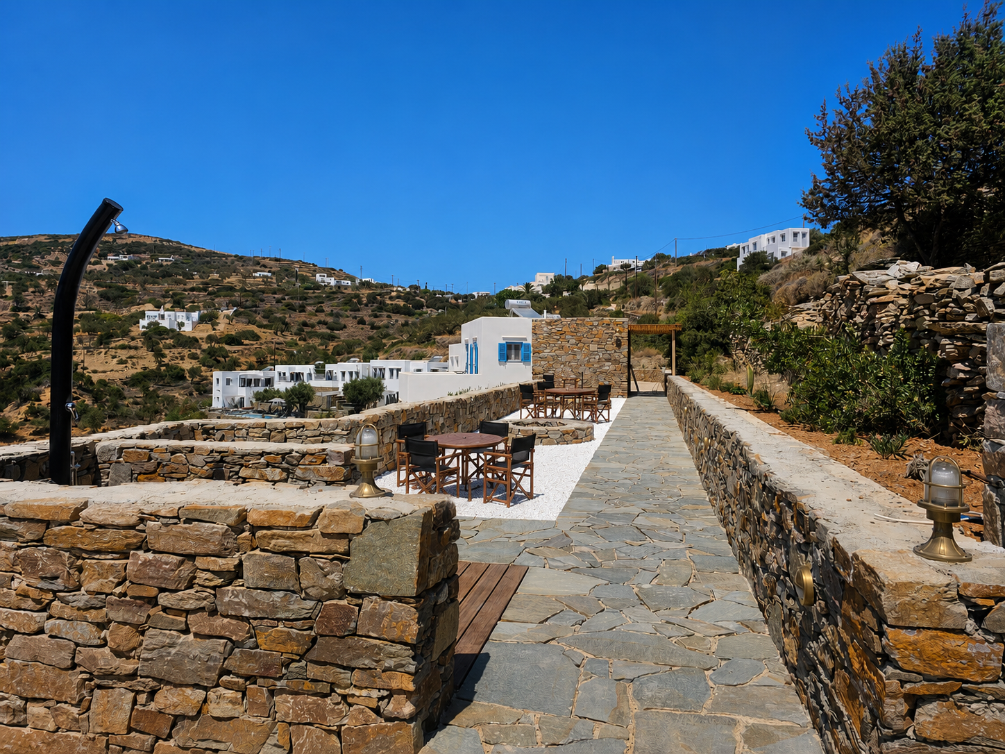 Stone walkway beside a low wall, leading to a white building under a bright blue sky in a rocky hillside setting