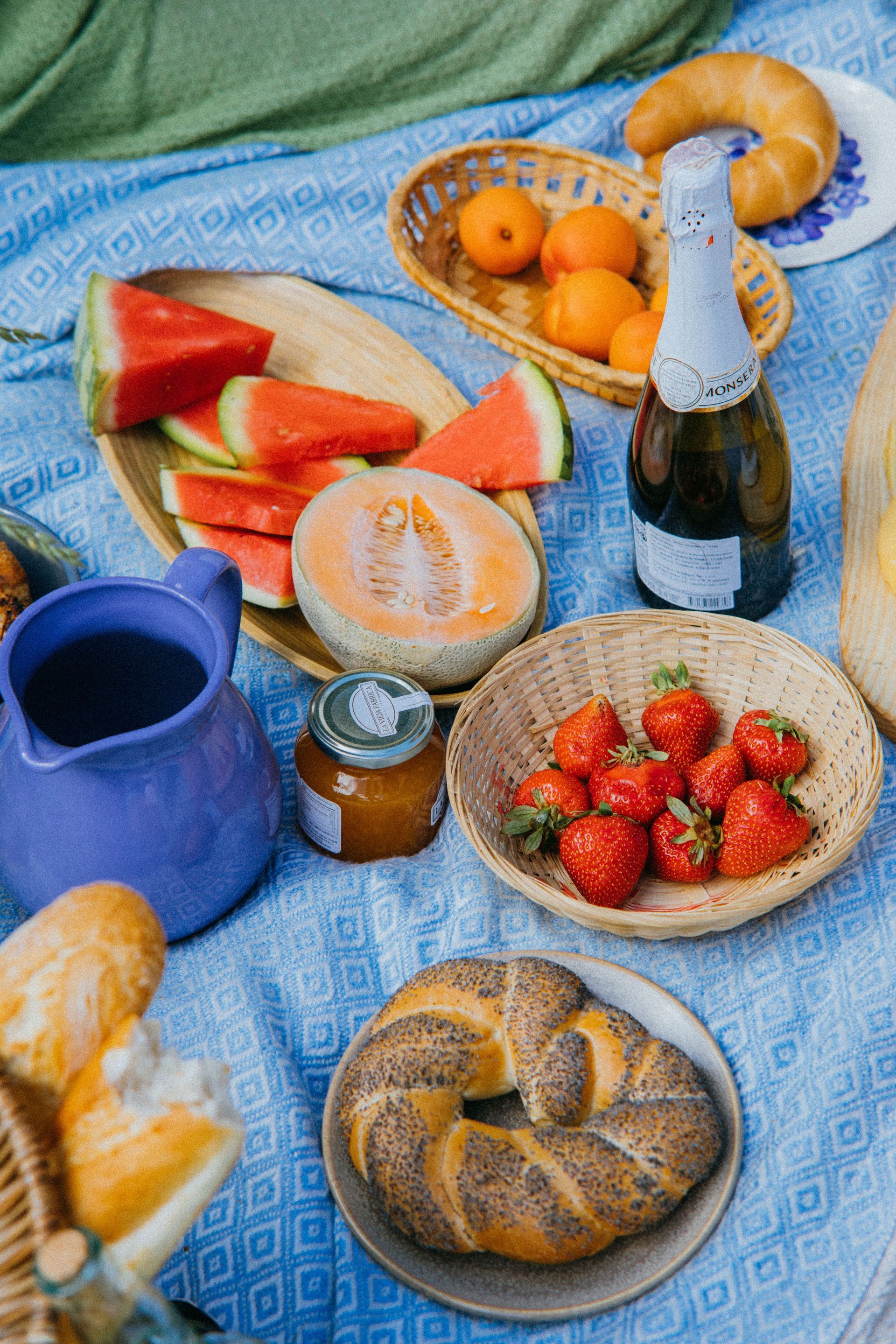 Picnic spread on a blue blanket with watermelon, strawberries, pastries, citrus, and a bottle of sparkling drink