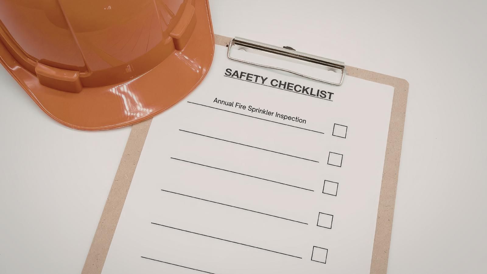 Safety checklist clipboard beside an orange hard hat on a white table.
