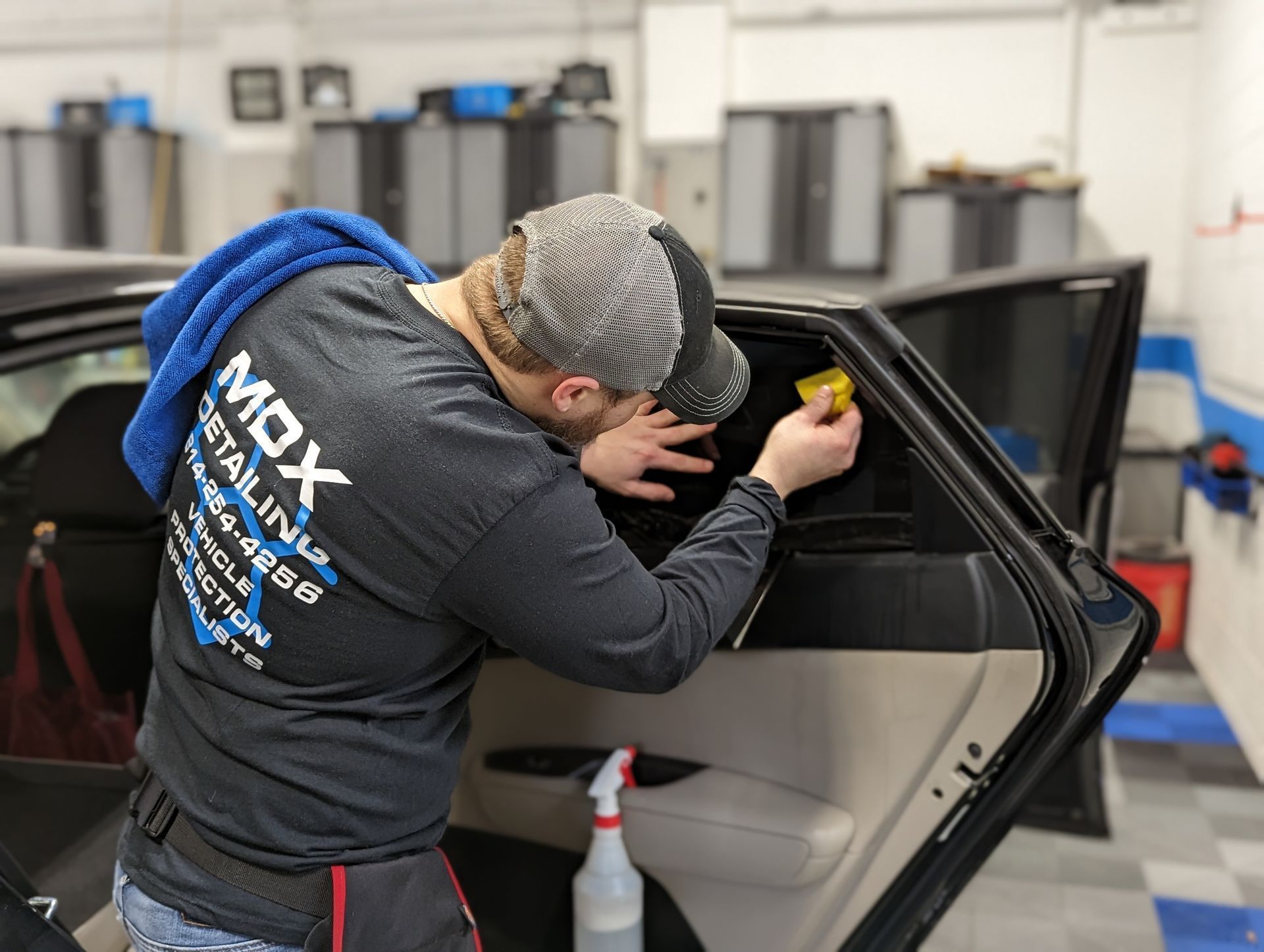 A man applying tint to a car window in a garage. He wears a hat and shirt for MDX Detailing. Window tint Johnstown PA